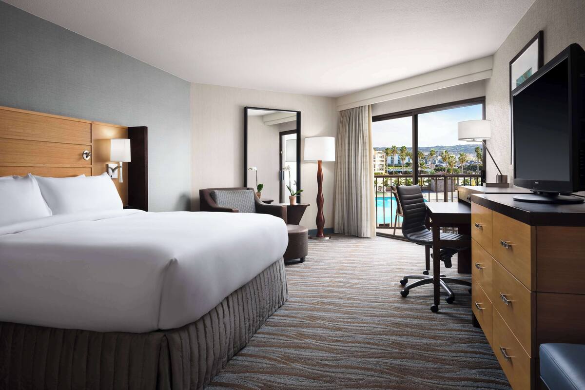 Une chambre d’hôtel avec un grand lit, un bureau avec une chaise, une télévision et une vue sur une piscine et des immeubles.