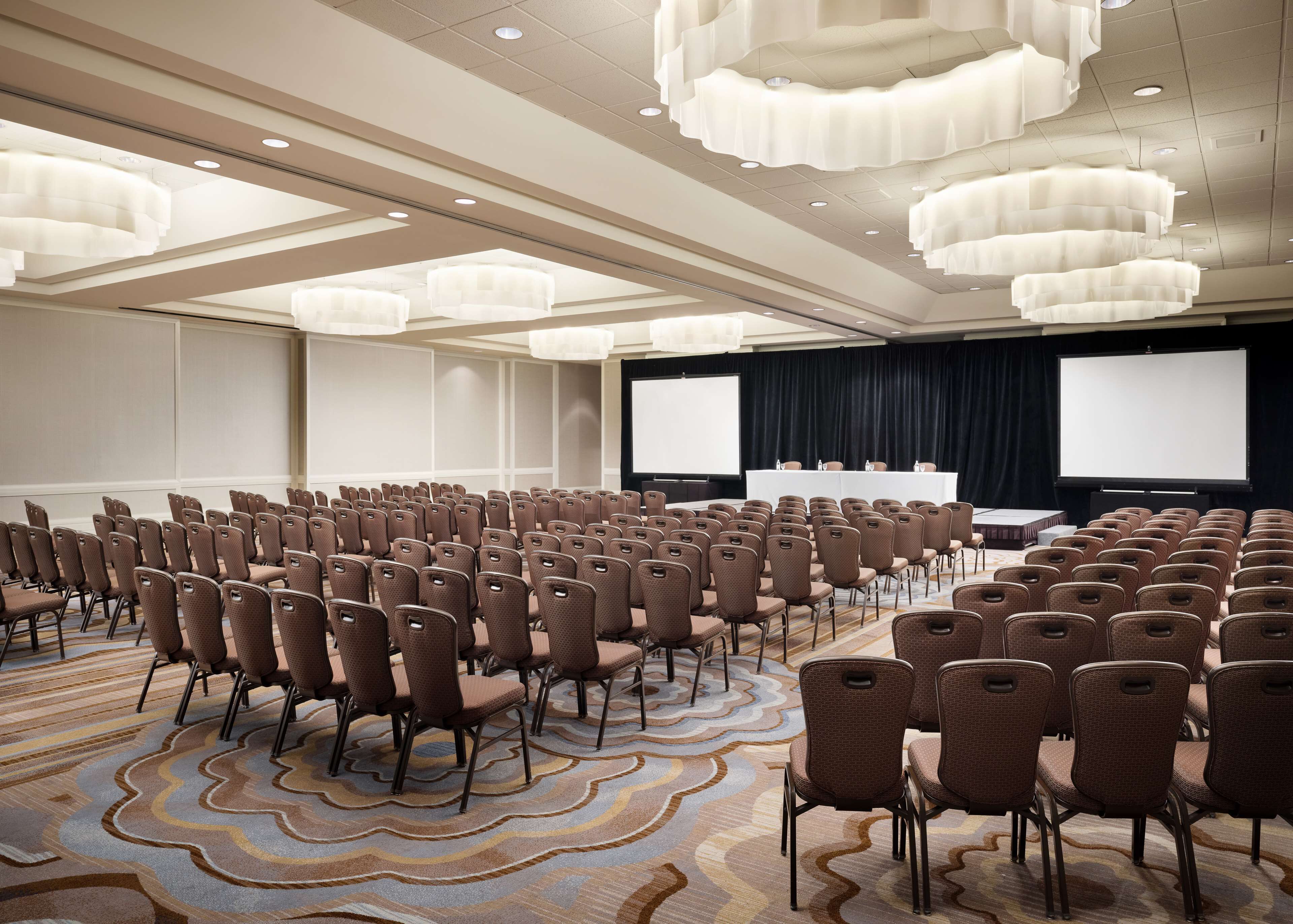 Un salón de conferencias con filas de sillas marrones, una pantalla grande y un escenario.