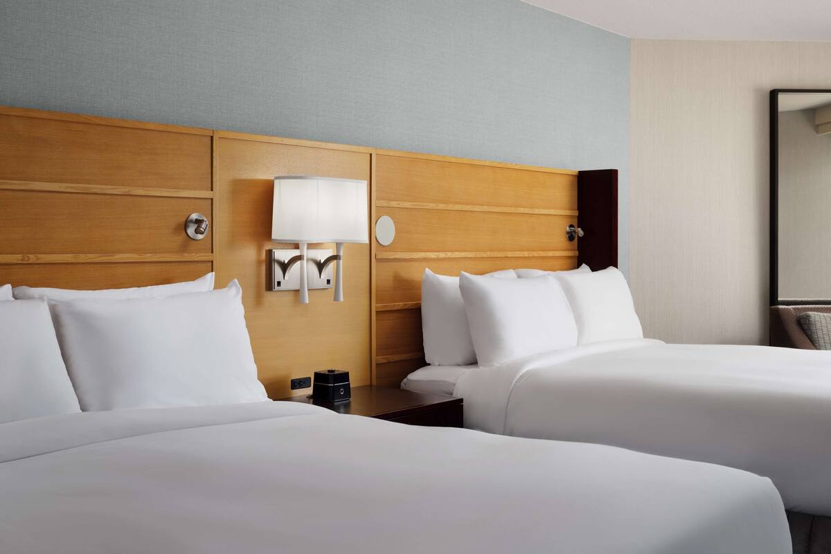 Une chambre d’hôtel avec deux lits, une tête de lit en bois, une lampe fixée au mur et une table de chevet avec un téléphone et une tasse.