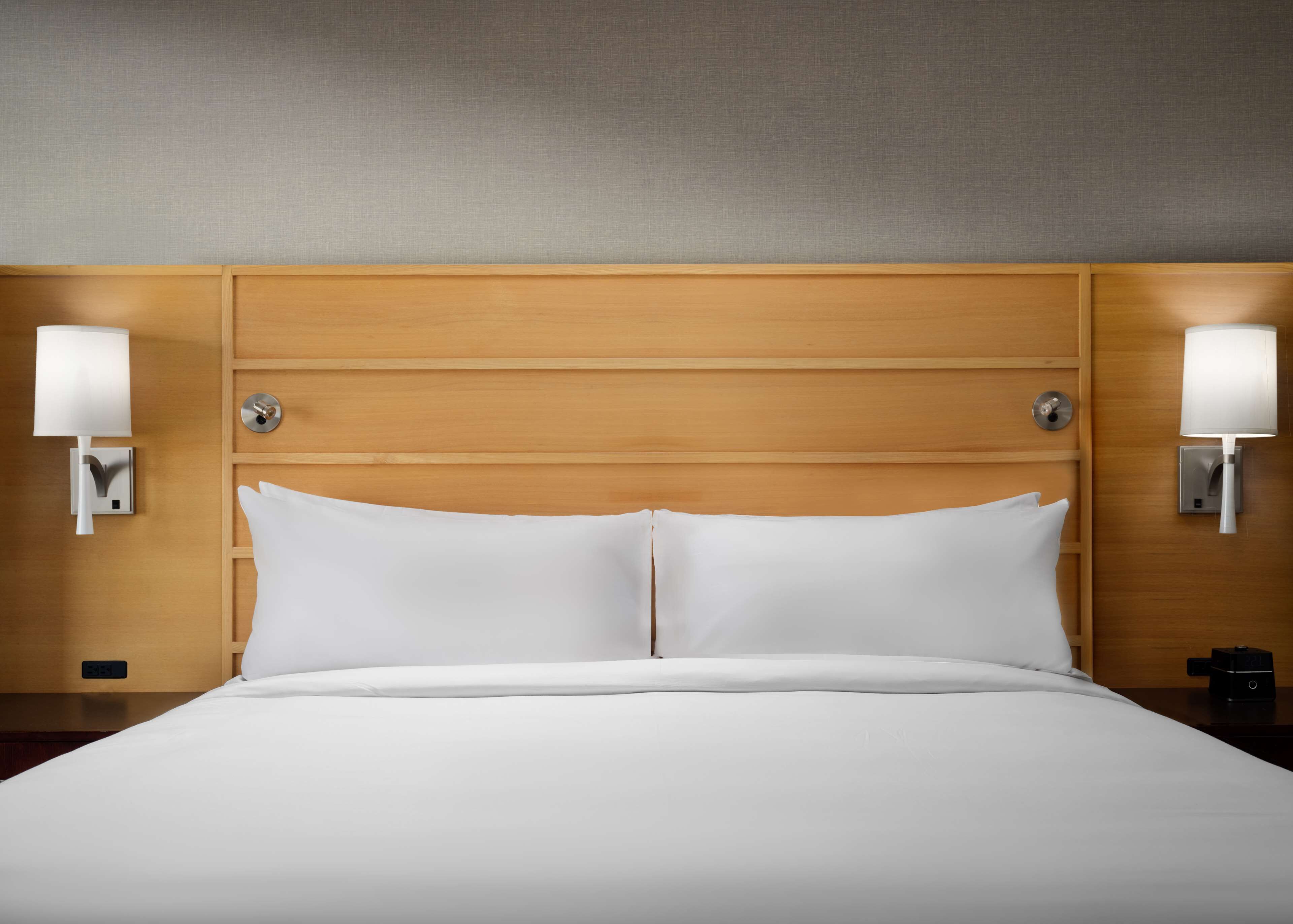 Une chambre d’hôtel avec une tête de lit en bois, deux lampes de chevet et une horloge numérique sur la table de chevet.