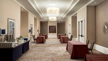 A hotel lobby with a long reception desk, multiple tables, and chairs, a patterned carpet, and a modern chandelier.