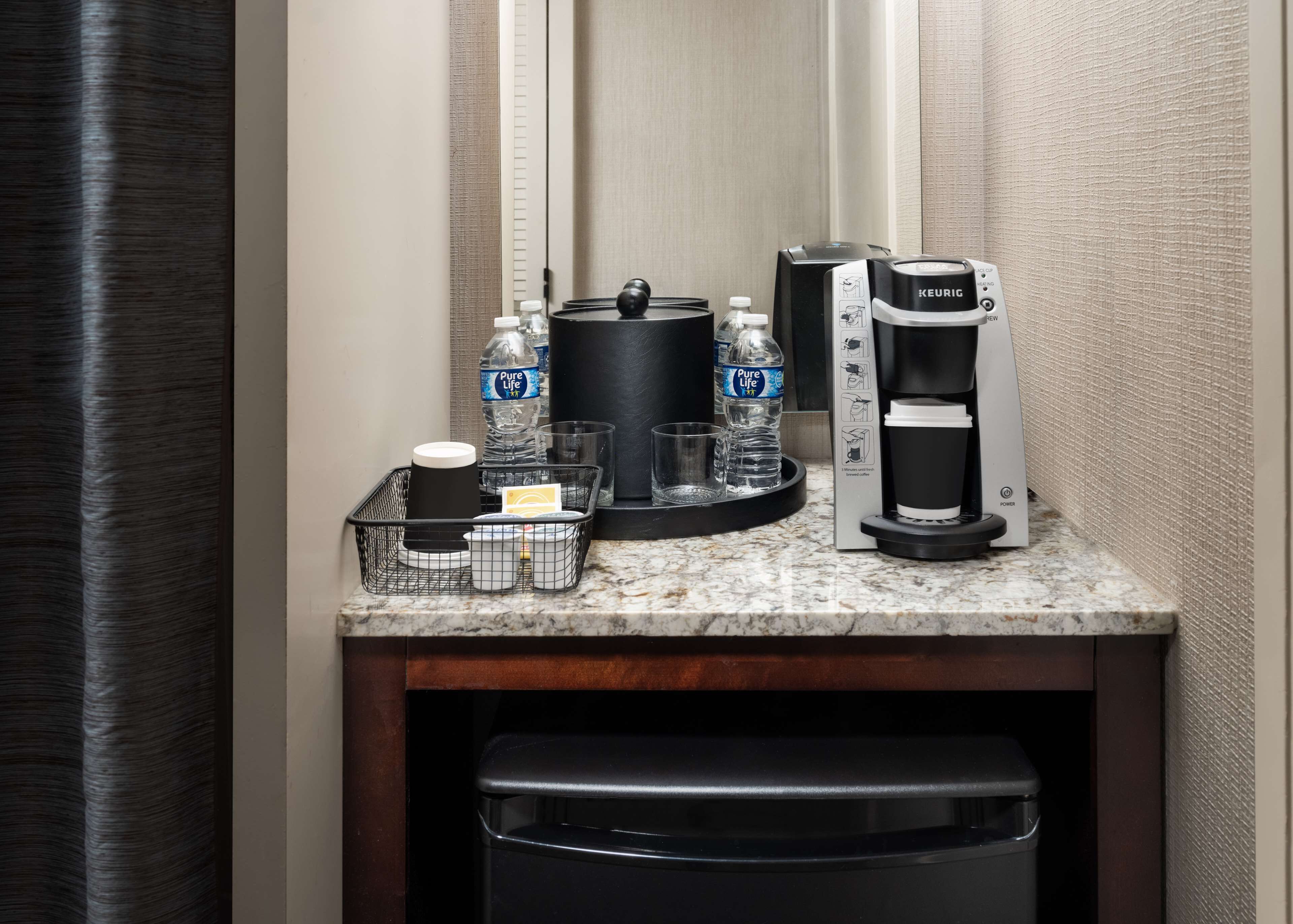 Ein Hotel-Kaffee-Bereich mit einer Keurig-Kaffeemaschine, zwei Wasserflaschen, einer Kaffeetasse und einem Mülleimer.