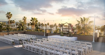A modern hotel room with a grey sofa, a matching armchair, a wooden partition, a small table with a lamp, and a view of the outdoors through large windows. at Sonesta Redondo Beach & Marina