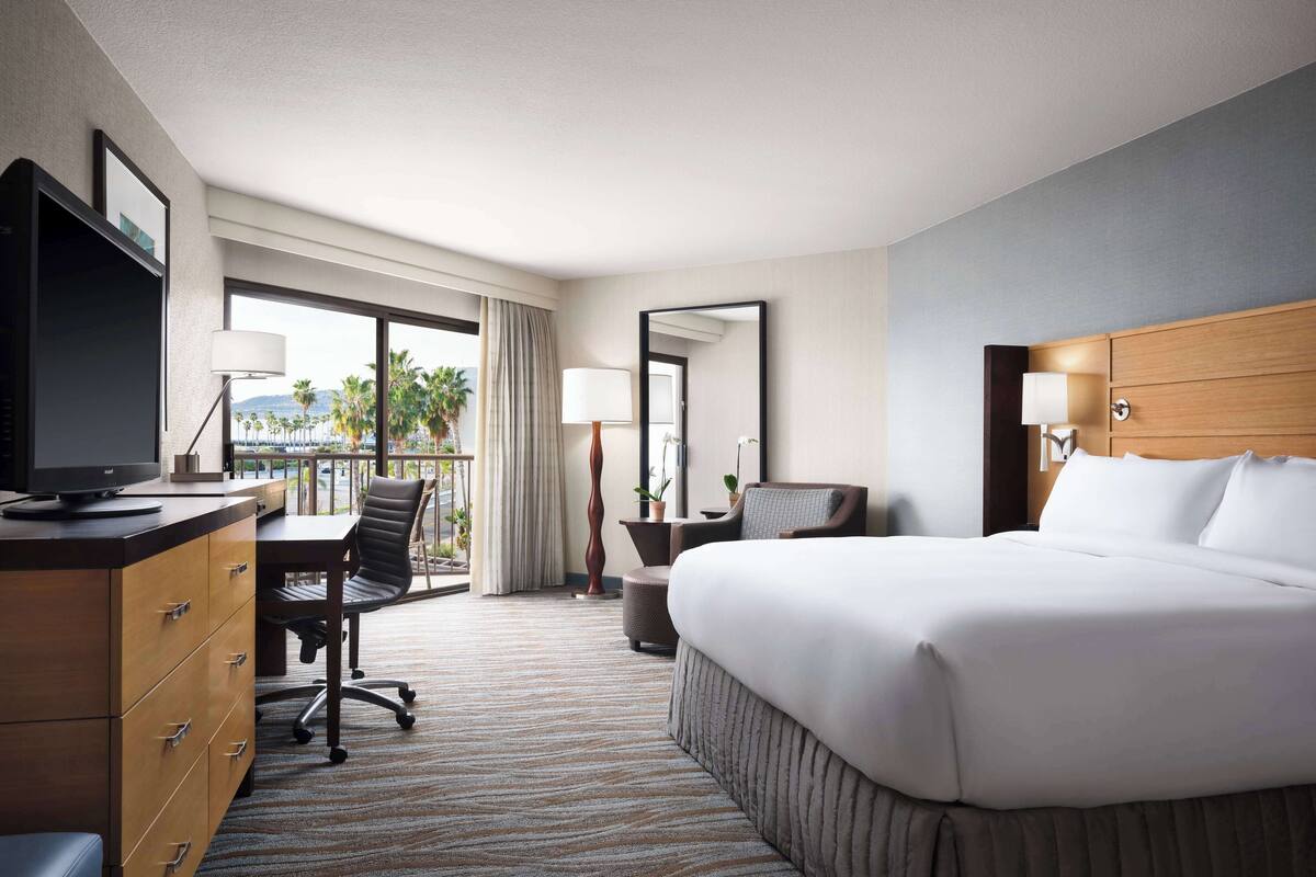 Une chambre d’hôtel avec un grand lit, un bureau avec un ordinateur, une chaise, une petite table avec une lampe et une fenêtre donnant sur des palmiers.