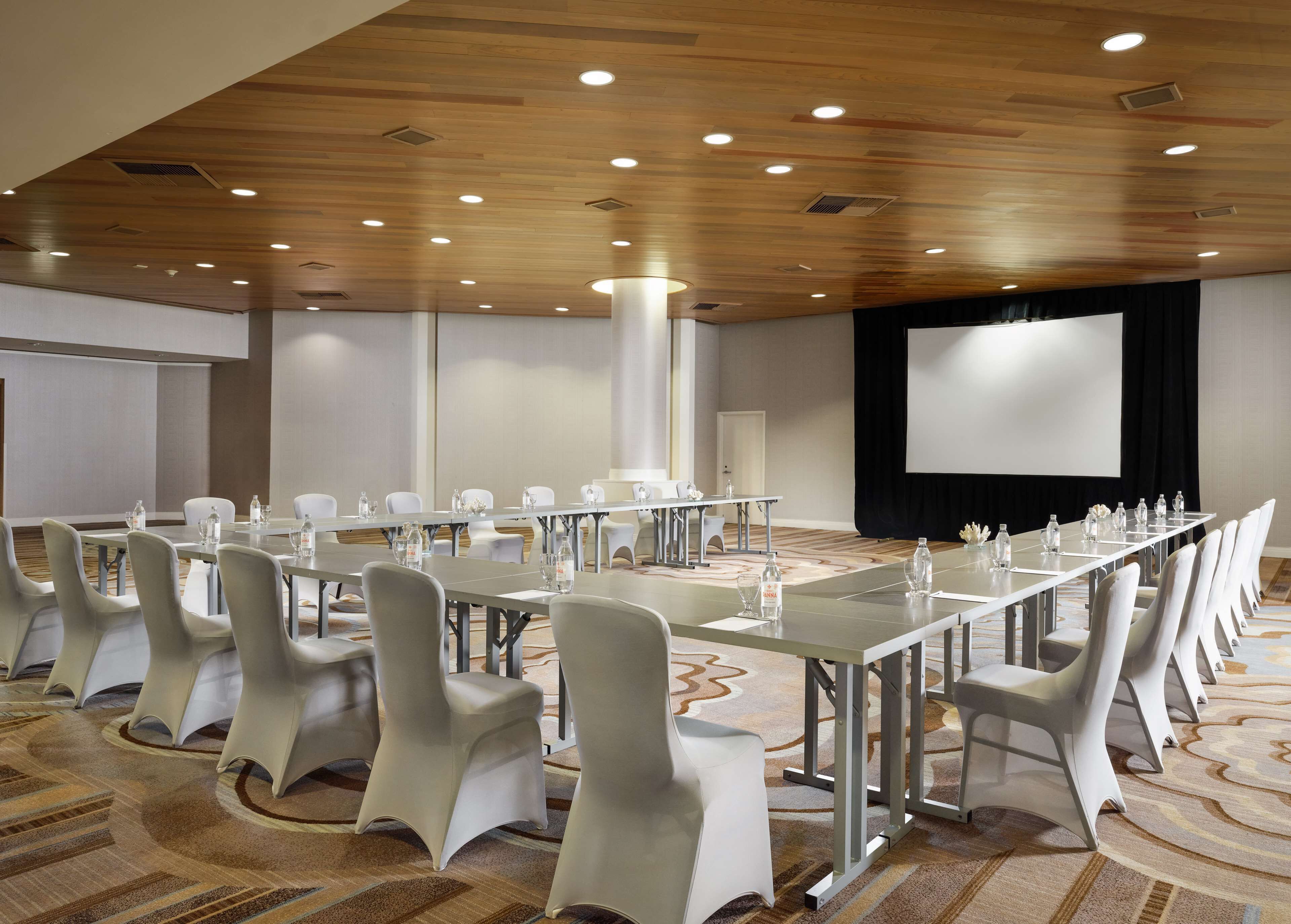 Ein Konferenzraum mit langem Tisch, weißen Stühlen, einer Projektionsleinwand und Teppichboden.