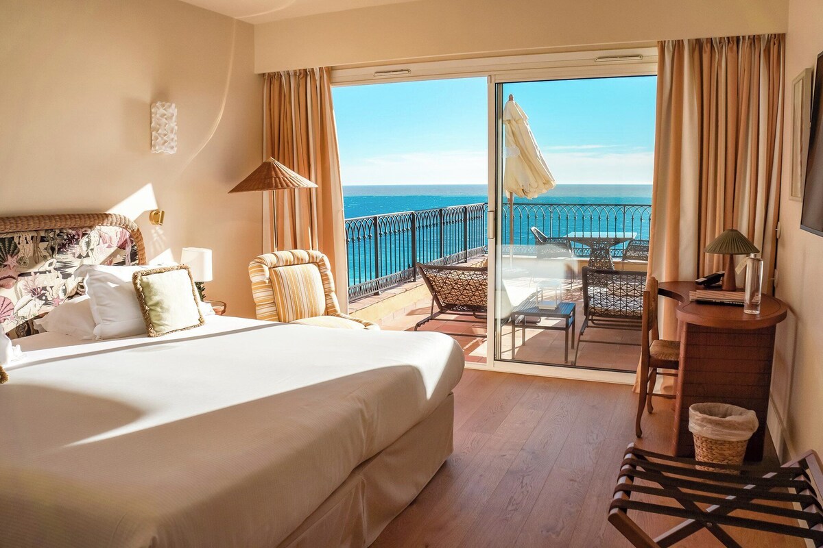 A hotel room with a large bed, a balcony with a view of the sea, and a small table with a lamp.