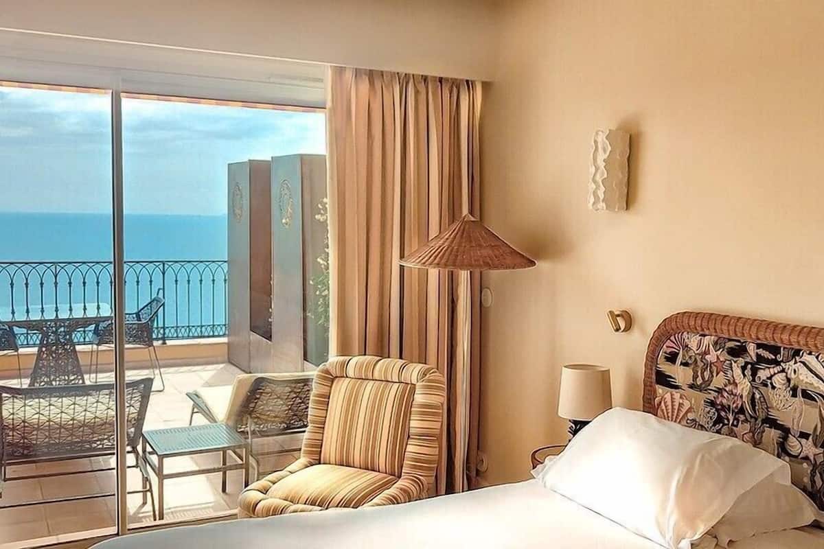 A hotel room with a bed, a chair, a table, a lamp, and a view of the sea.