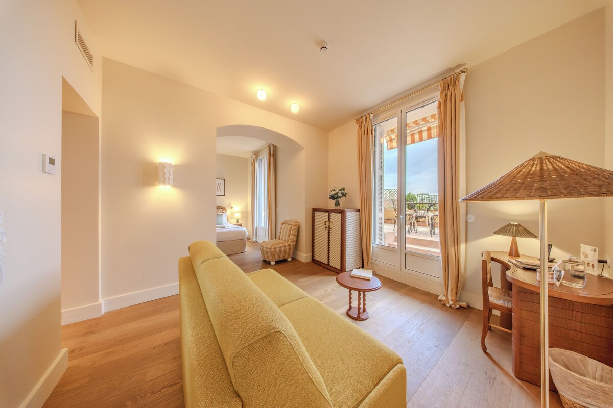 A hotel room with a sofa, a small table, a chair, a cabinet, and a view of the outside.