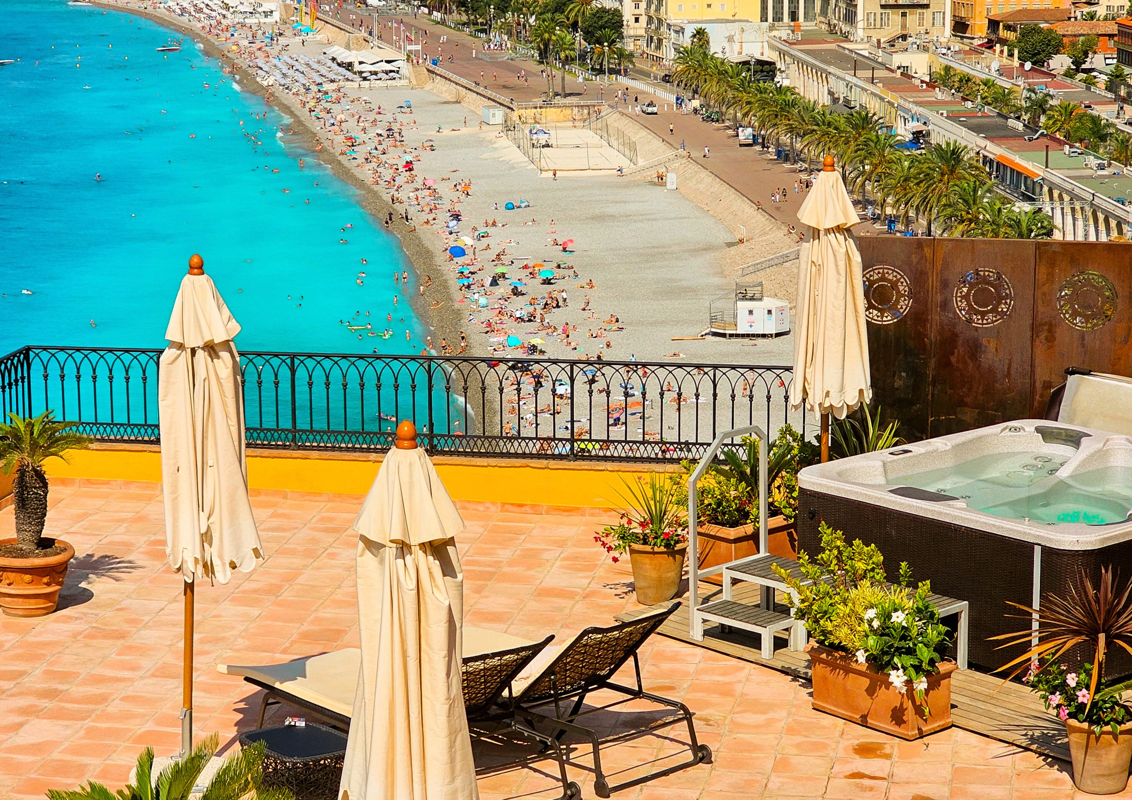 A rooftop terrace with outdoor seating, a hot tub, and a view of a busy beach with people and umbrellas.
