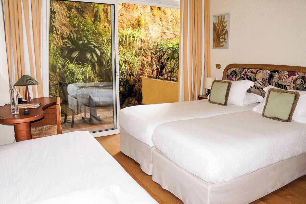 A hotel room with a large bed, a small table with a lamp, and a view of greenery through a sliding door.
