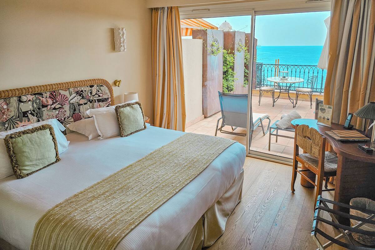 A hotel room with a bed, a desk, and a balcony with a view of the sea.