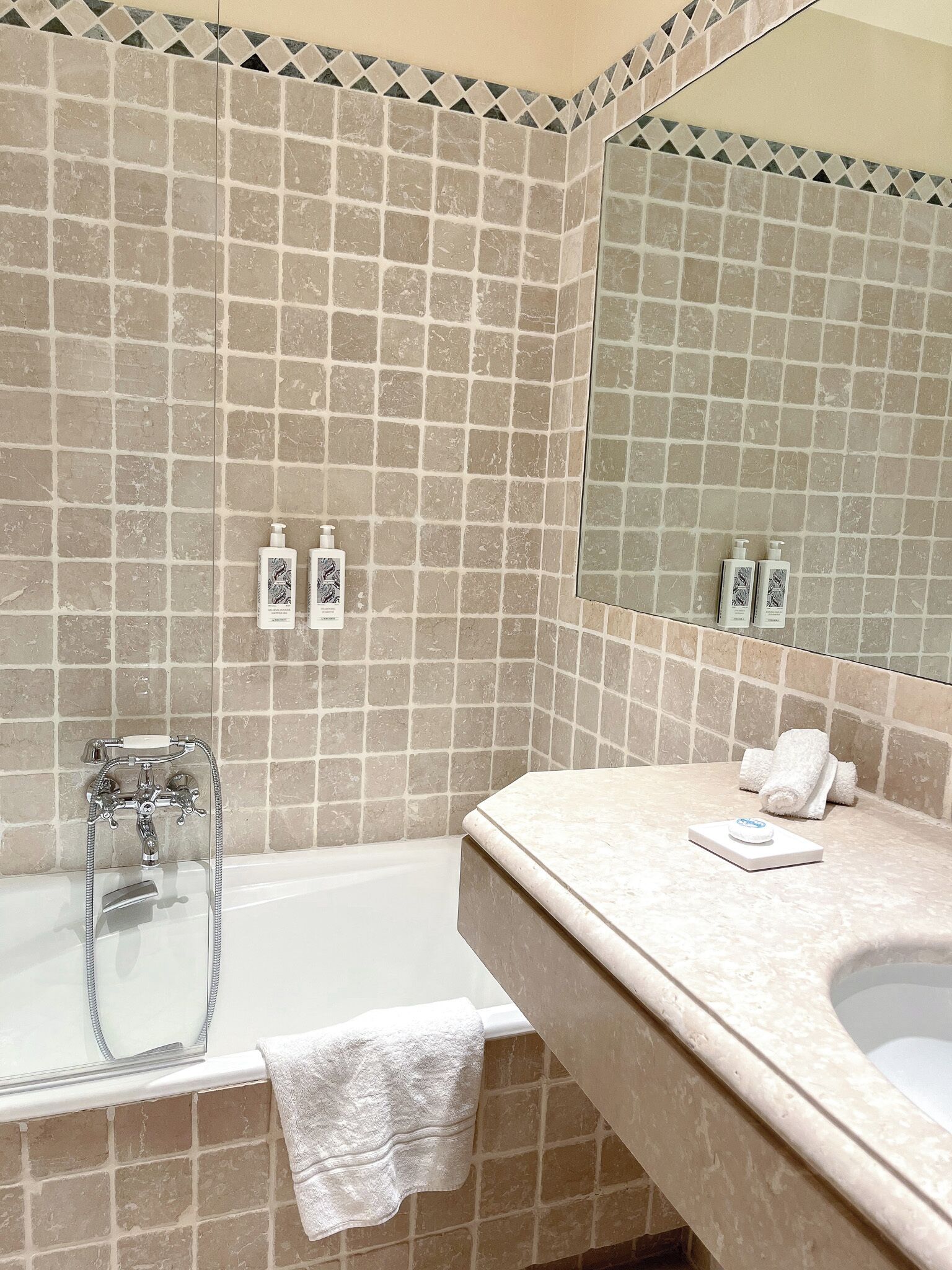 Een badkamer met een badkuip, een wastafel en een spiegel. Op het aanrecht staan twee flessen shampoo en een handdoek.