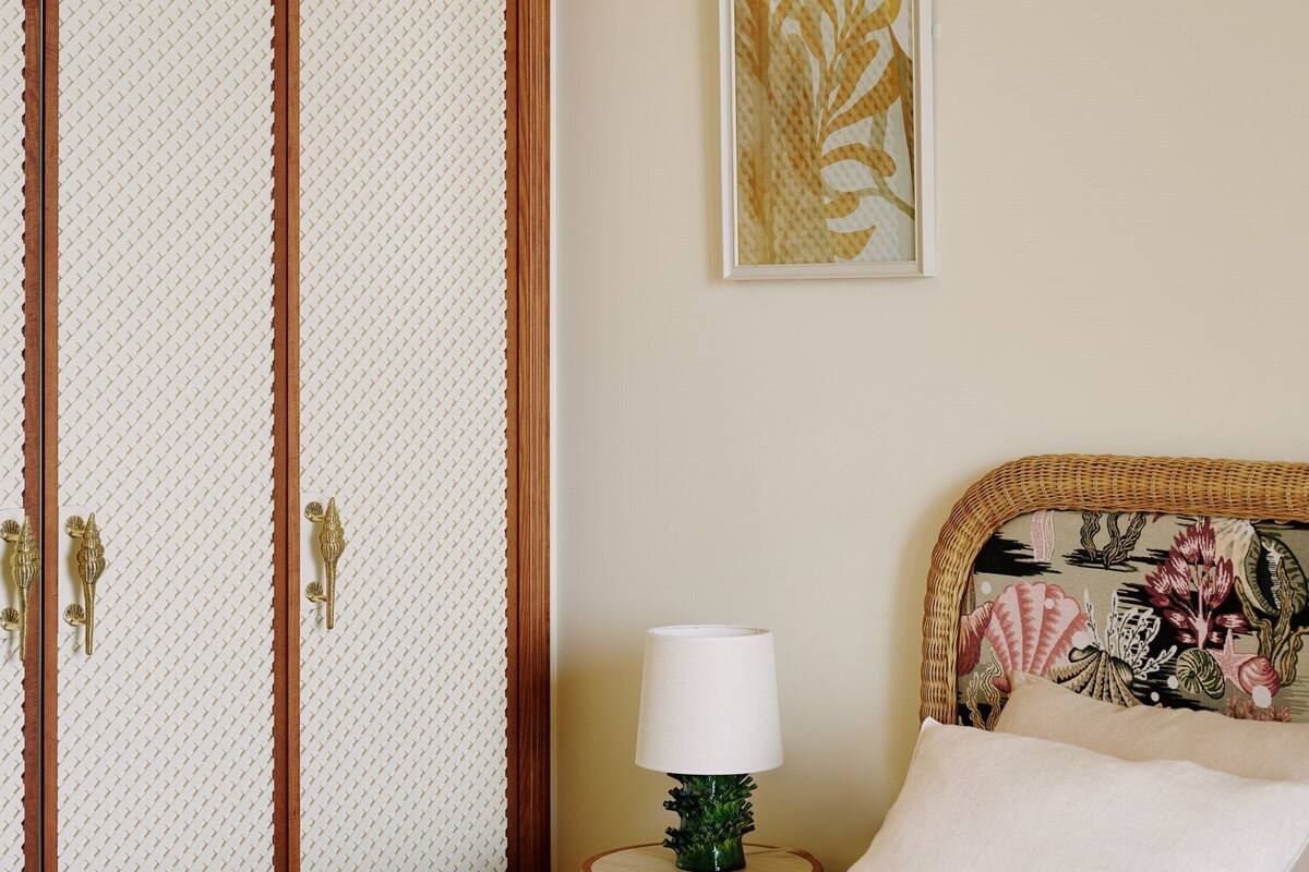 A bedroom with a bed, a nightstand with a lamp, a decorative headboard, a wardrobe, and a framed artwork on the wall.