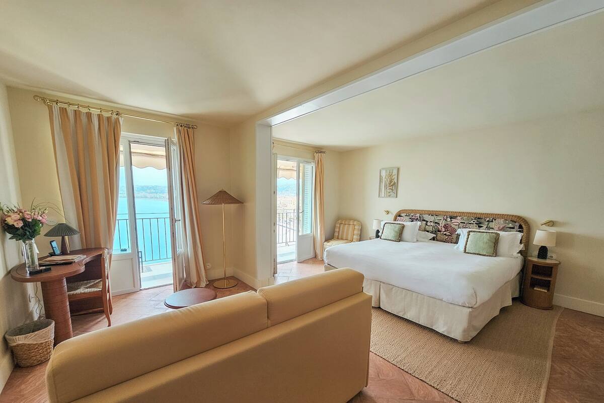 A hotel room with a bed, a sofa, a desk with a lamp, and a view of the sea.