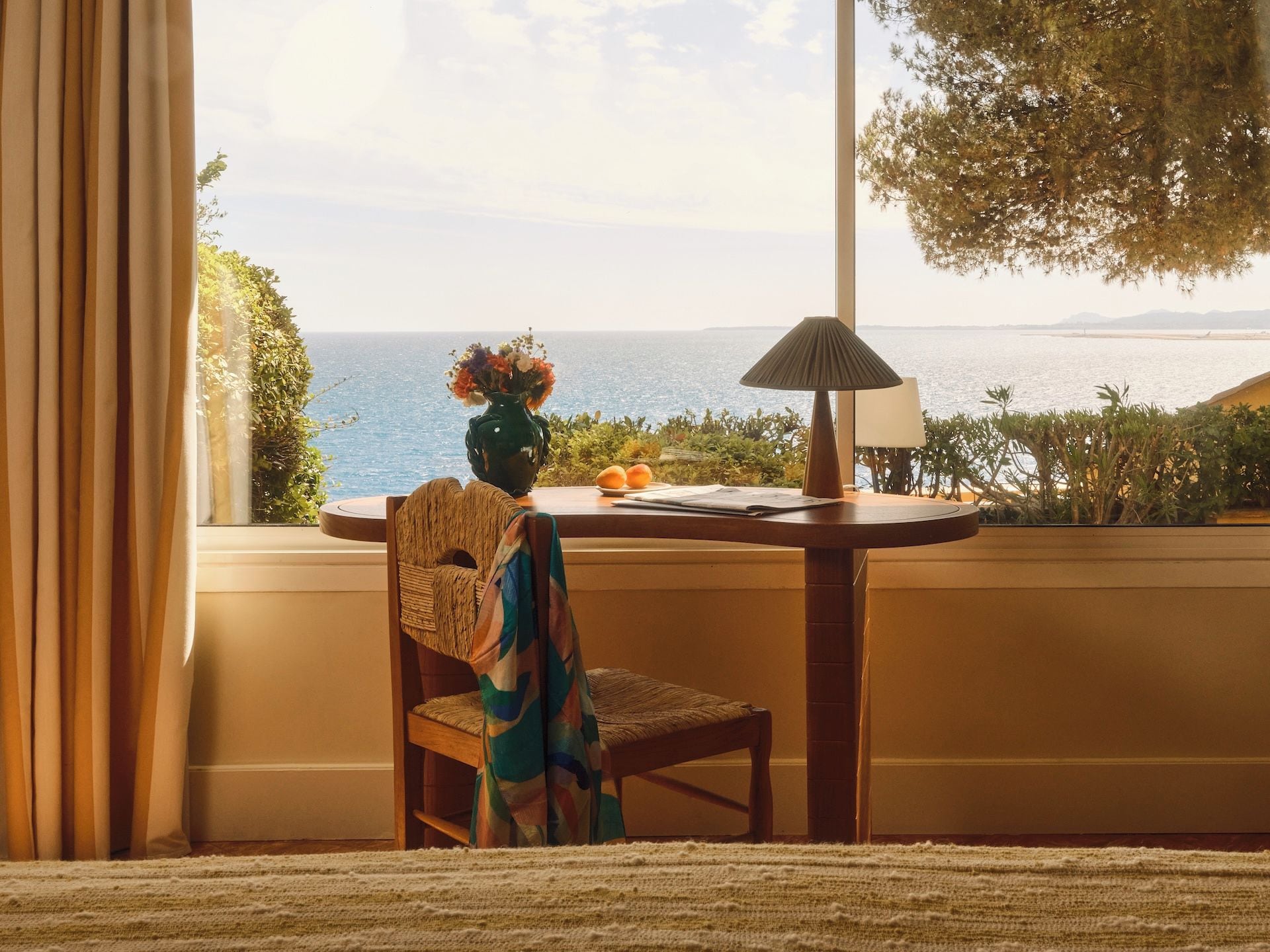 A room with a view of the sea, a table with a lamp, and a chair with a woven backrest.