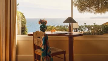 A room with a view of the sea, a table with a lamp, and a chair with a woven backrest.