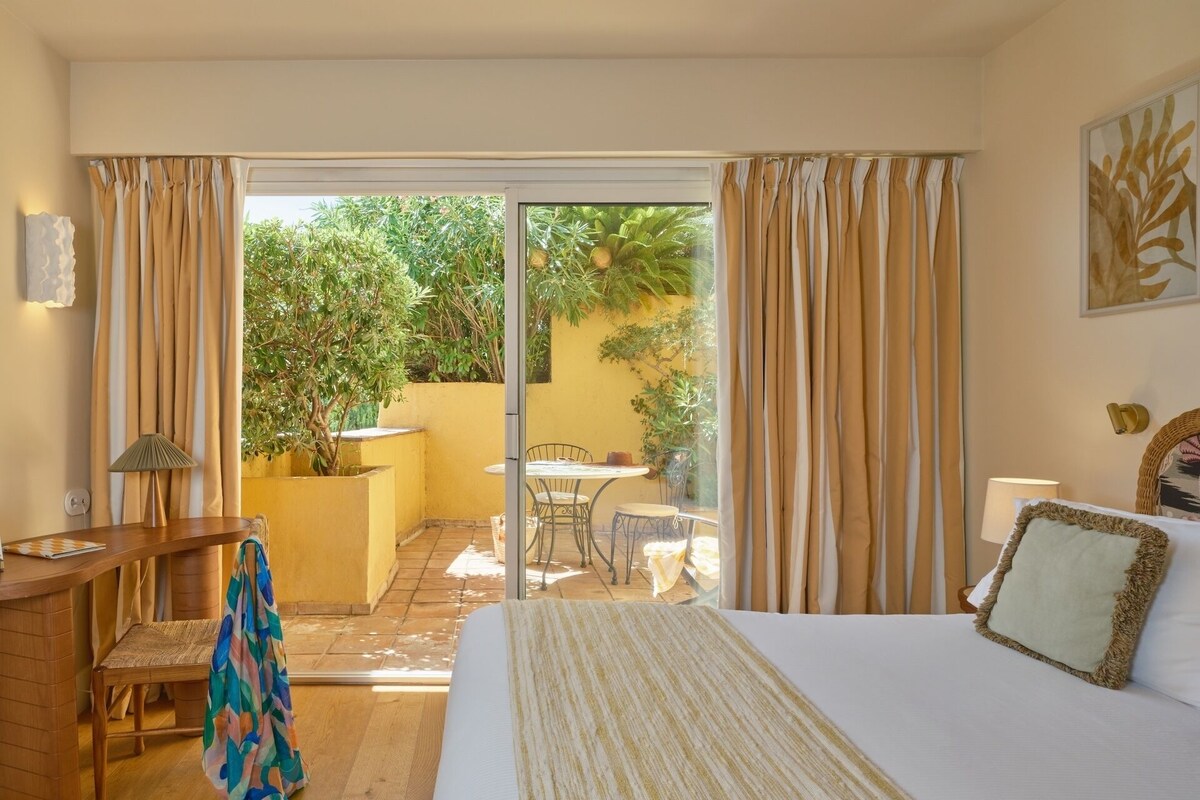 A hotel room with a bed, a desk, a chair, a small table, and a view of the garden through the sliding glass door.