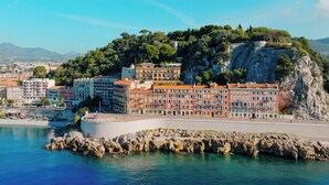 A coastal cityscape with a prominent building complex on a hill, a beach, and rocky shoreline. - Hôtel La Pérouse Nice (Nice)