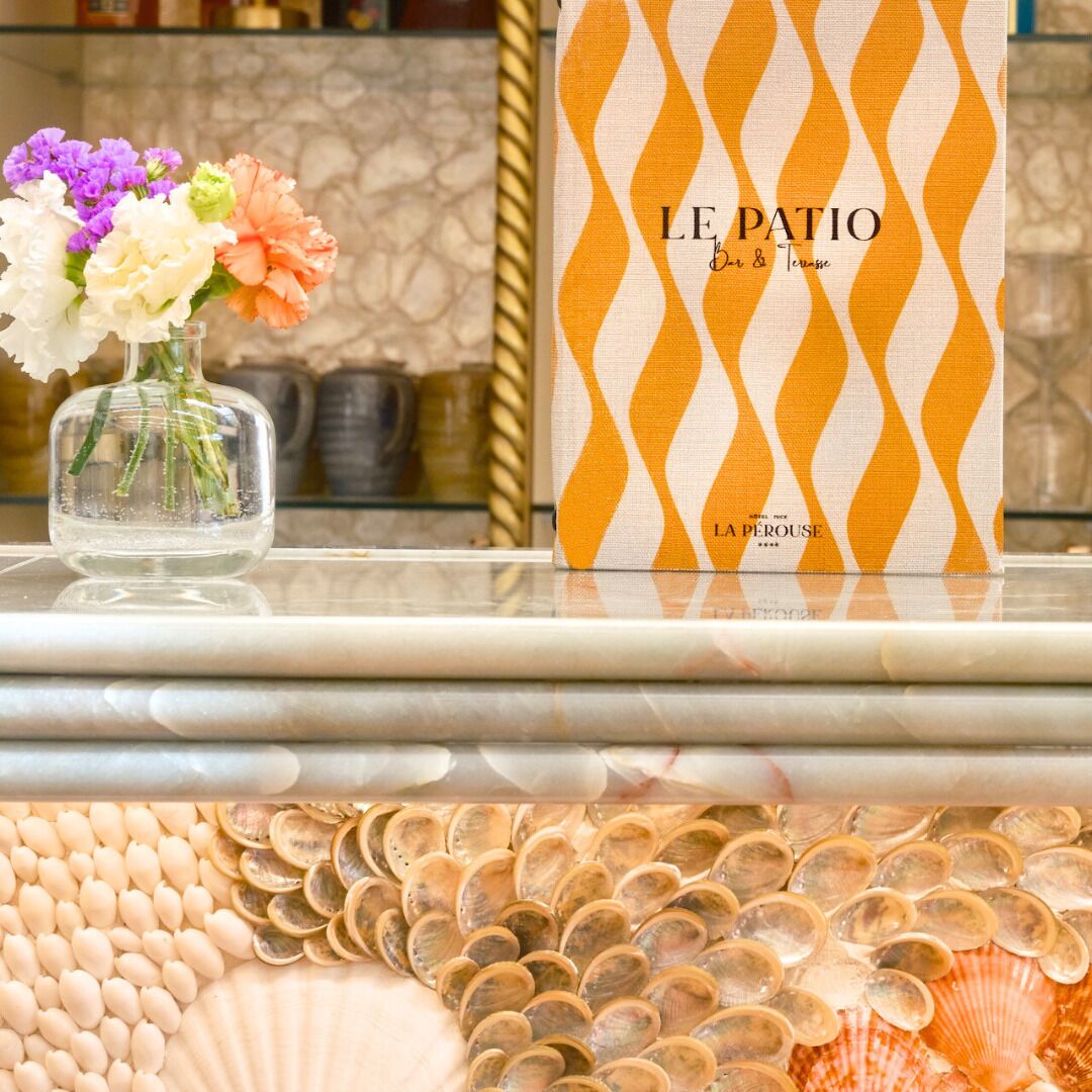 Un vaso di vetro trasparente con fiori e una scatola decorata con su scritto “LE PATIO bar & terrace” su una superficie riflettente.
