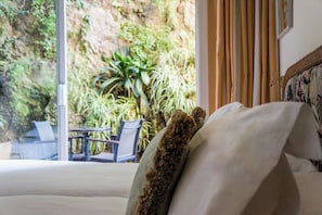 A hotel room with a large window, a bed with a white headboard, and a view of a lush garden outside.