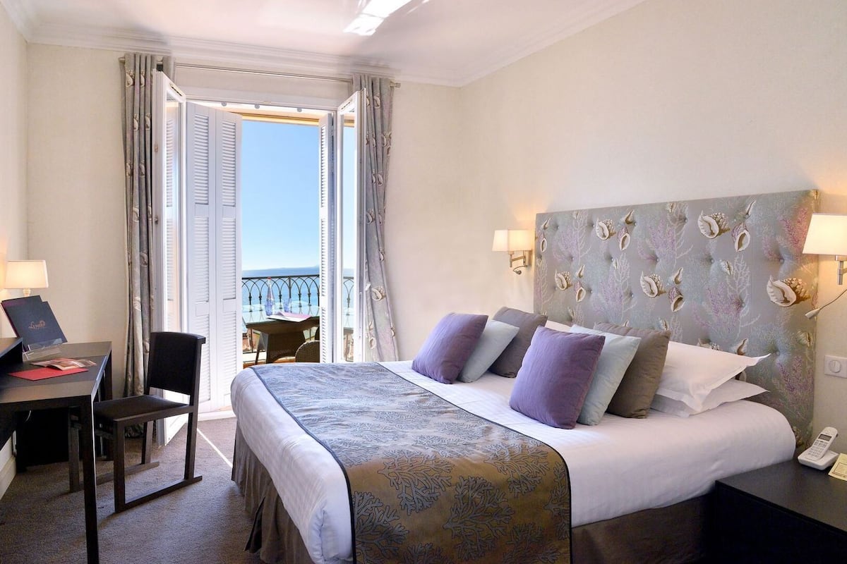 A hotel room with a bed, a desk, a chair, a nightstand, and a view of the sea through an open door.