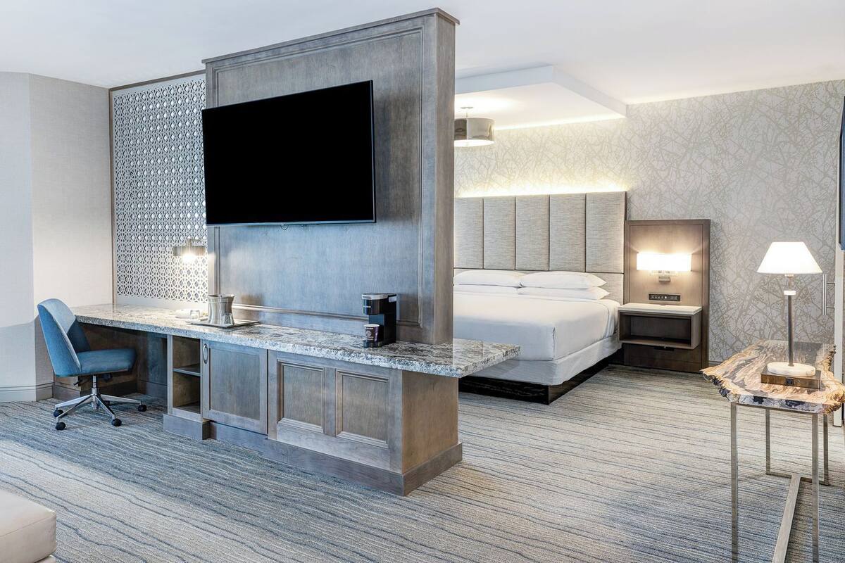 A hotel room with a bed, a desk with a flat-screen TV, a chair, a nightstand, and a lamp.