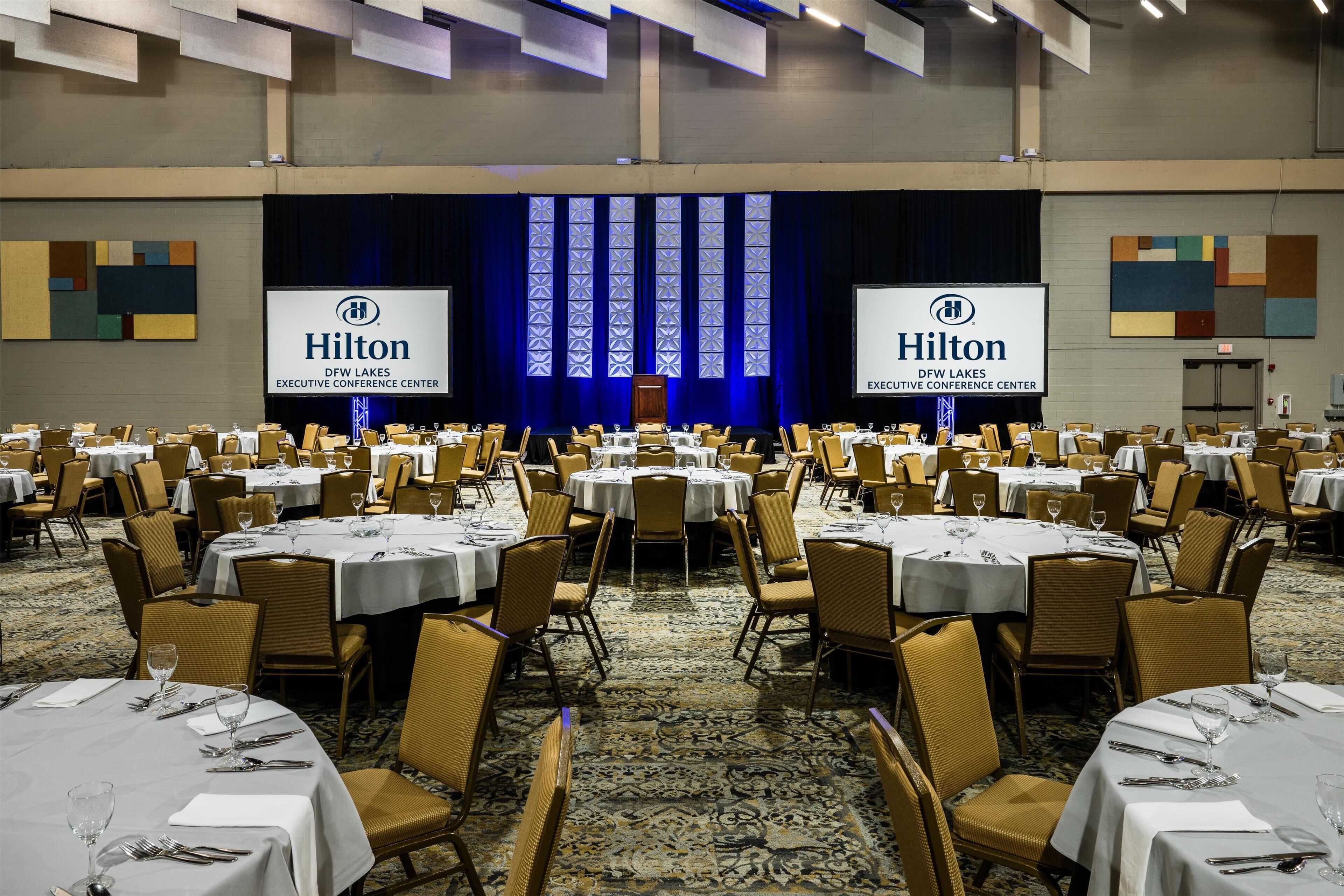 A large conference room with round tables and chairs, a stage with a blue curtain, and two Hilton Executive Conference Center signs.