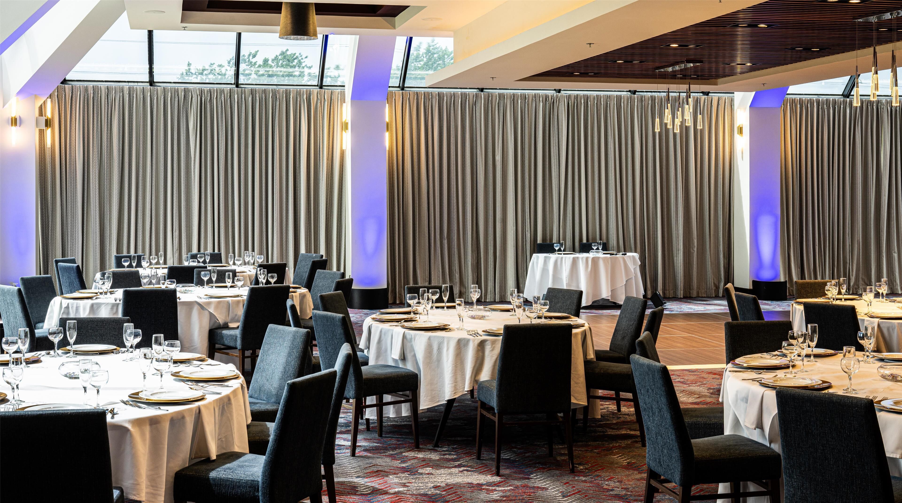 A banquet hall with round tables set for dining, large windows with curtains, and modern lighting fixtures.