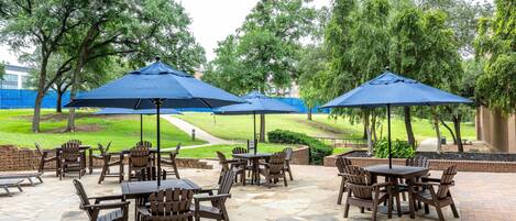 An outdoor patio area with multiple tables and chairs, surrounded by trees and covered by blue umbrellas.