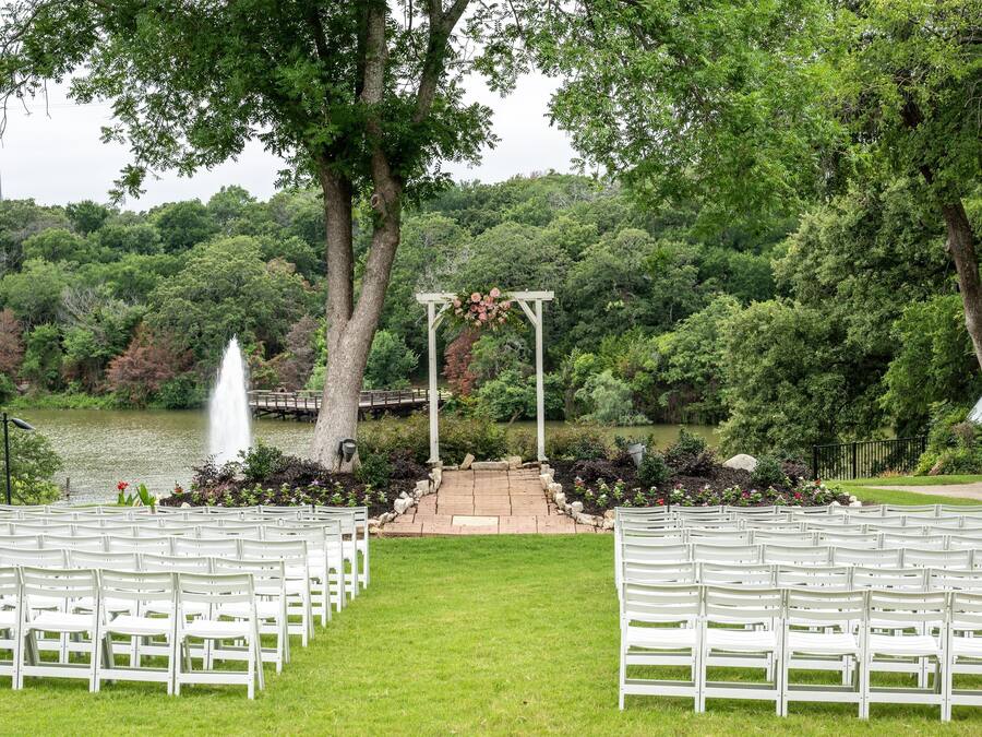 A wedding ceremony setup with white chairs arranged in rows, a wooden arch with flowers, and a fountain in the background.