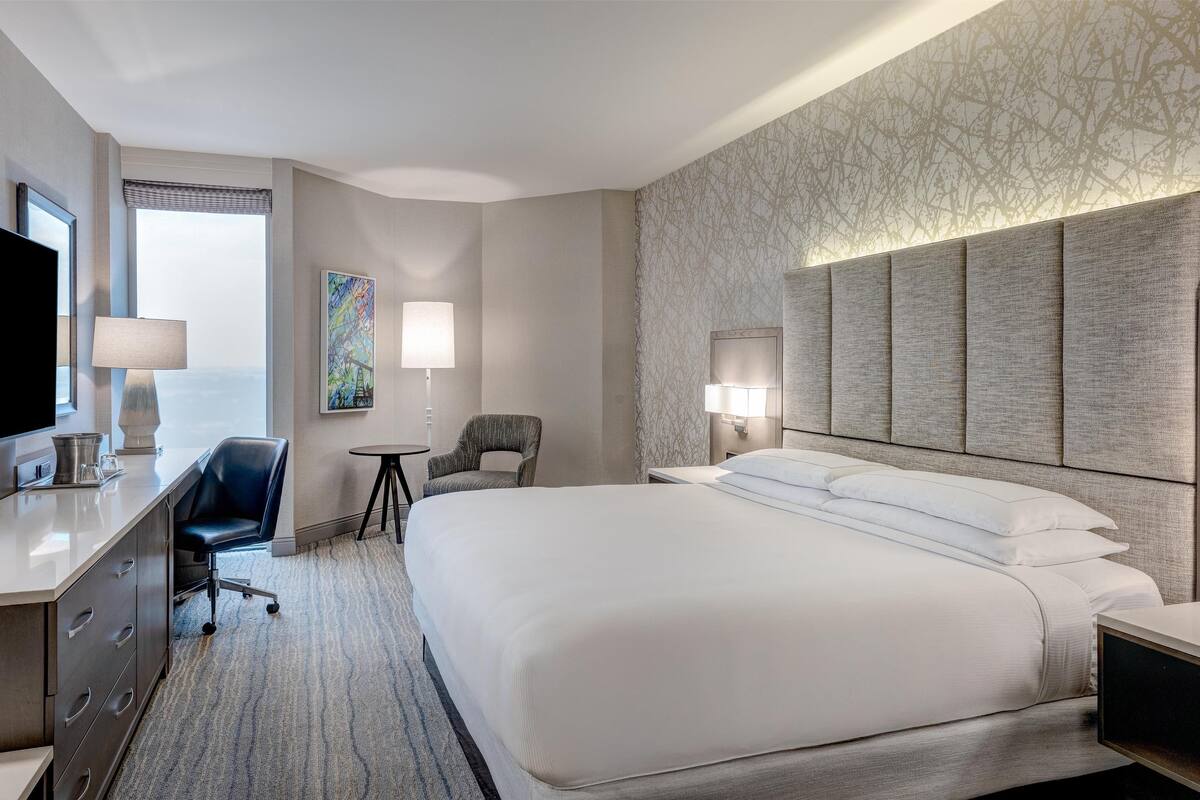 A hotel room with a large bed, a desk with a flat-screen TV, a chair, a small table, and a lamp.