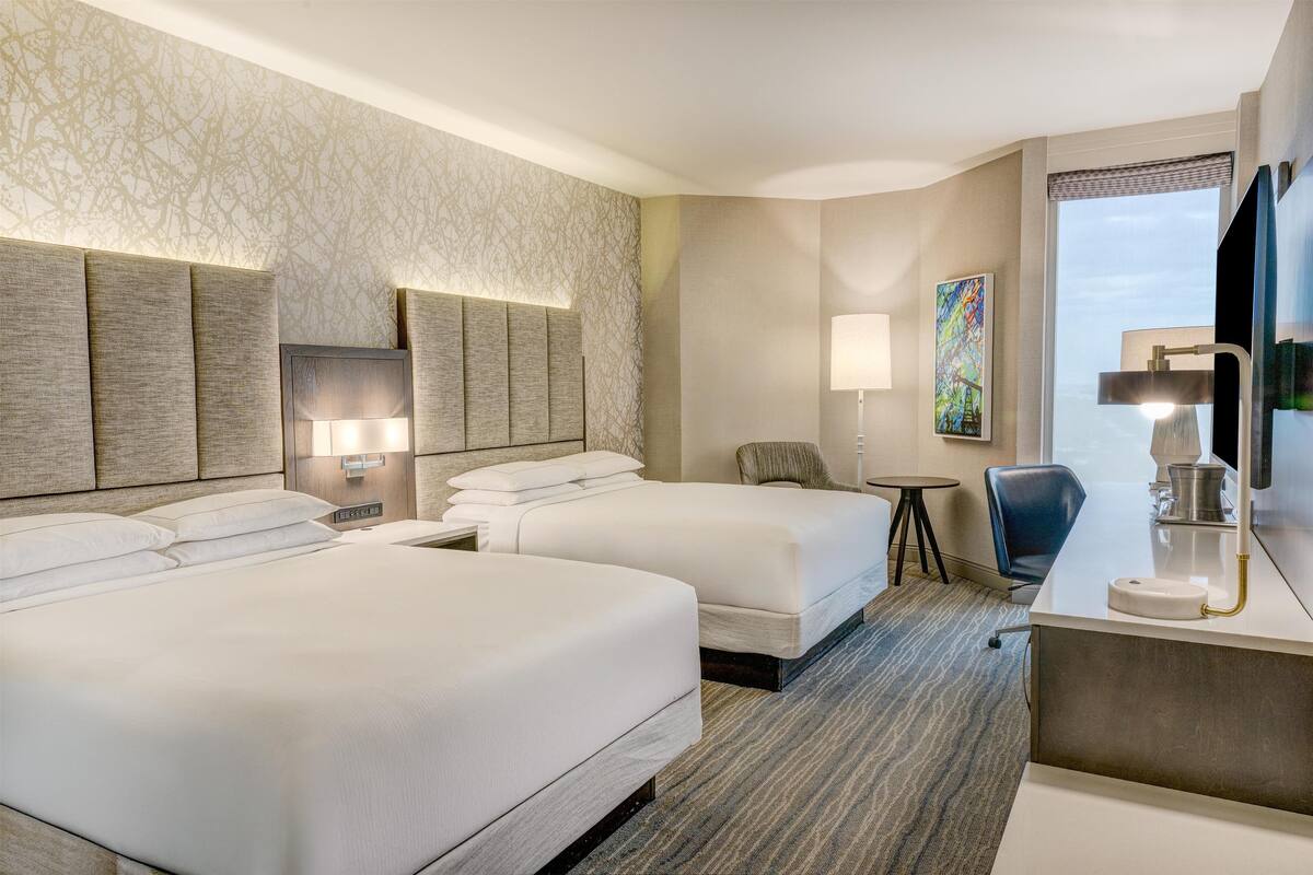 A hotel room with two beds, a desk, a chair, a small table, and a painting on the wall.