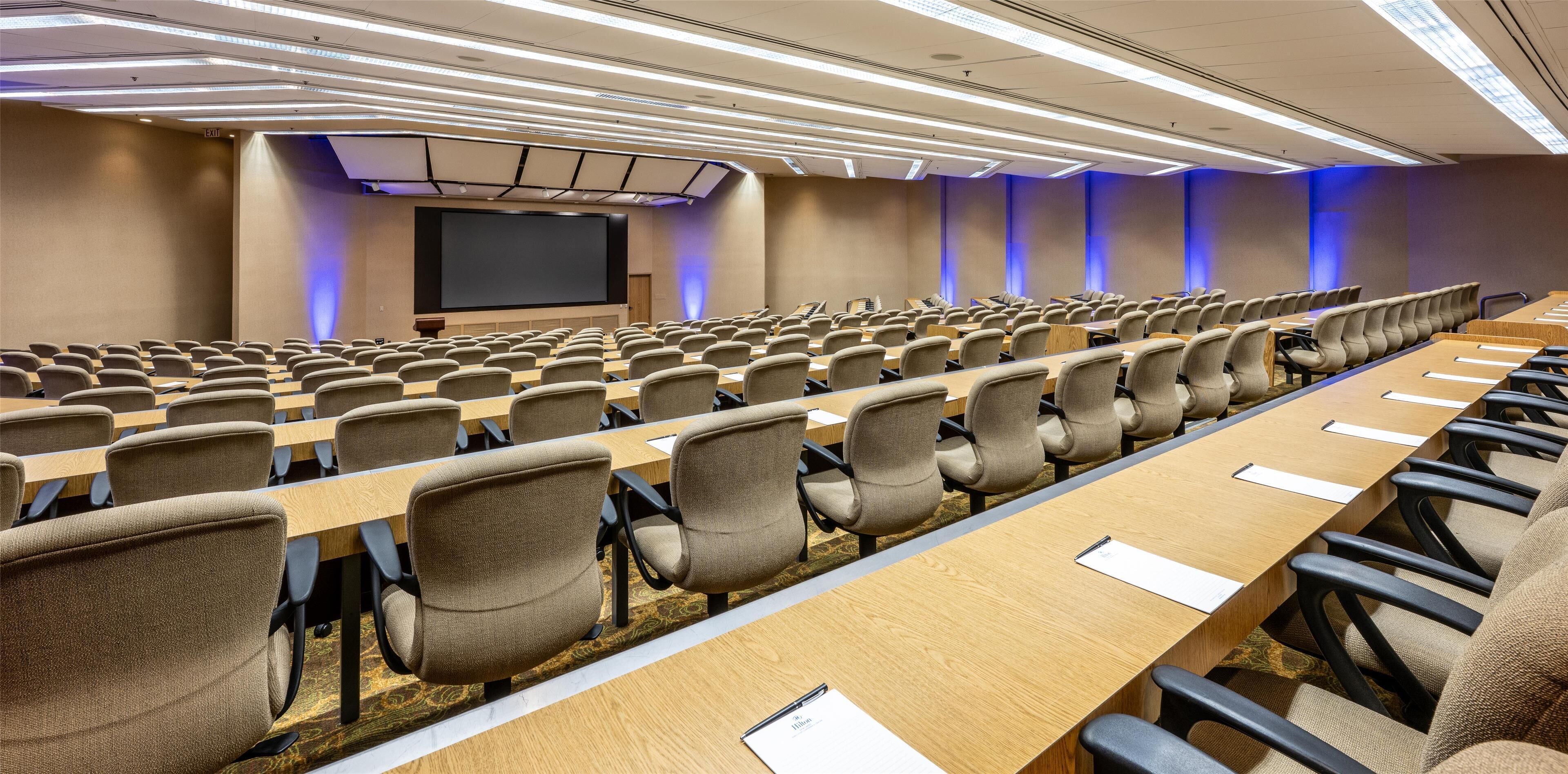 A conference room with rows of chairs and tables, a large screen, and blue ambient lighting.