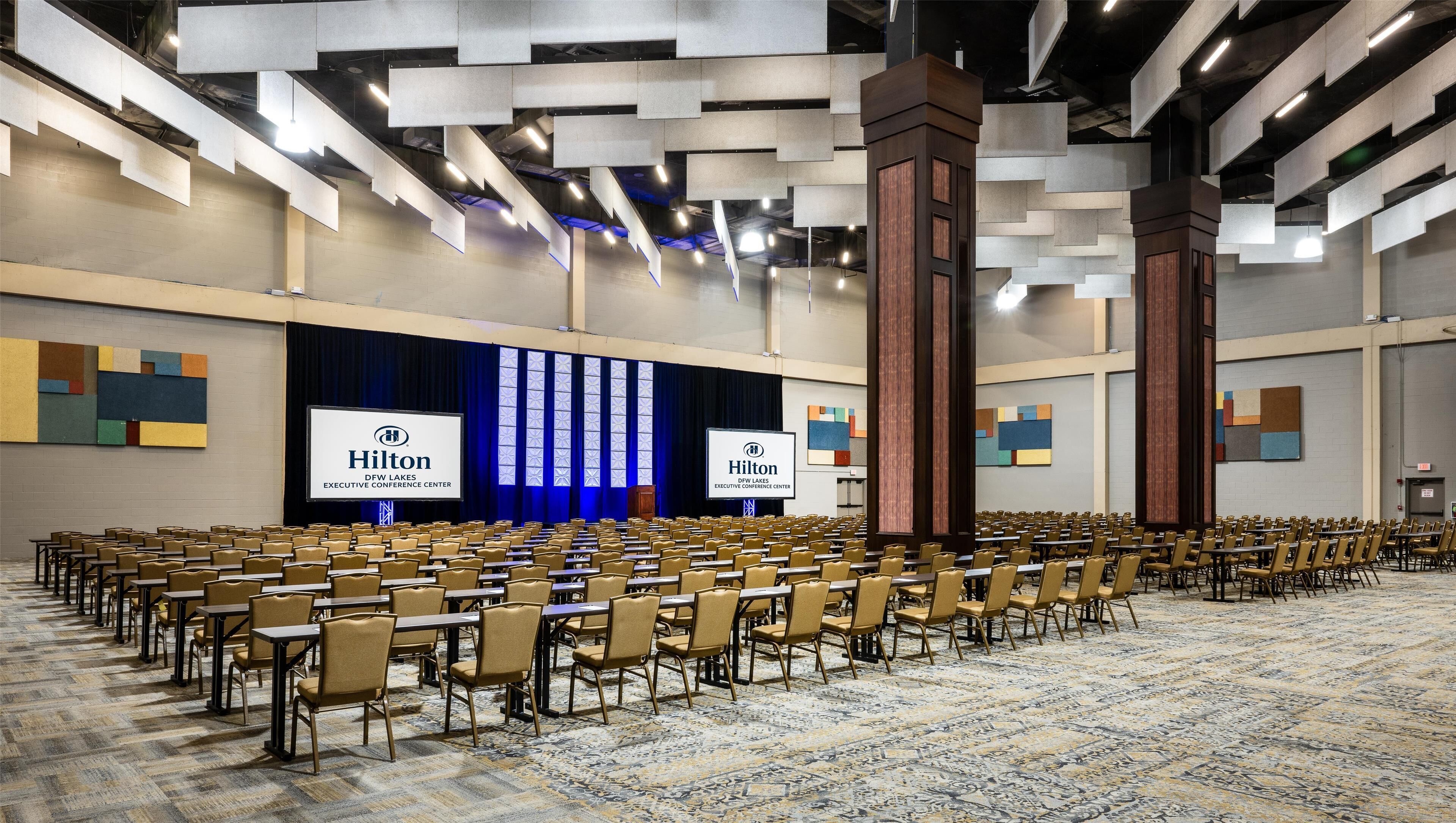 A large conference room with rows of tables and chairs, a stage with a Hilton logo, and a carpeted floor.