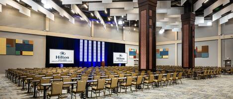 A large conference room with rows of tables and chairs, a stage with a Hilton logo, and a carpeted floor.