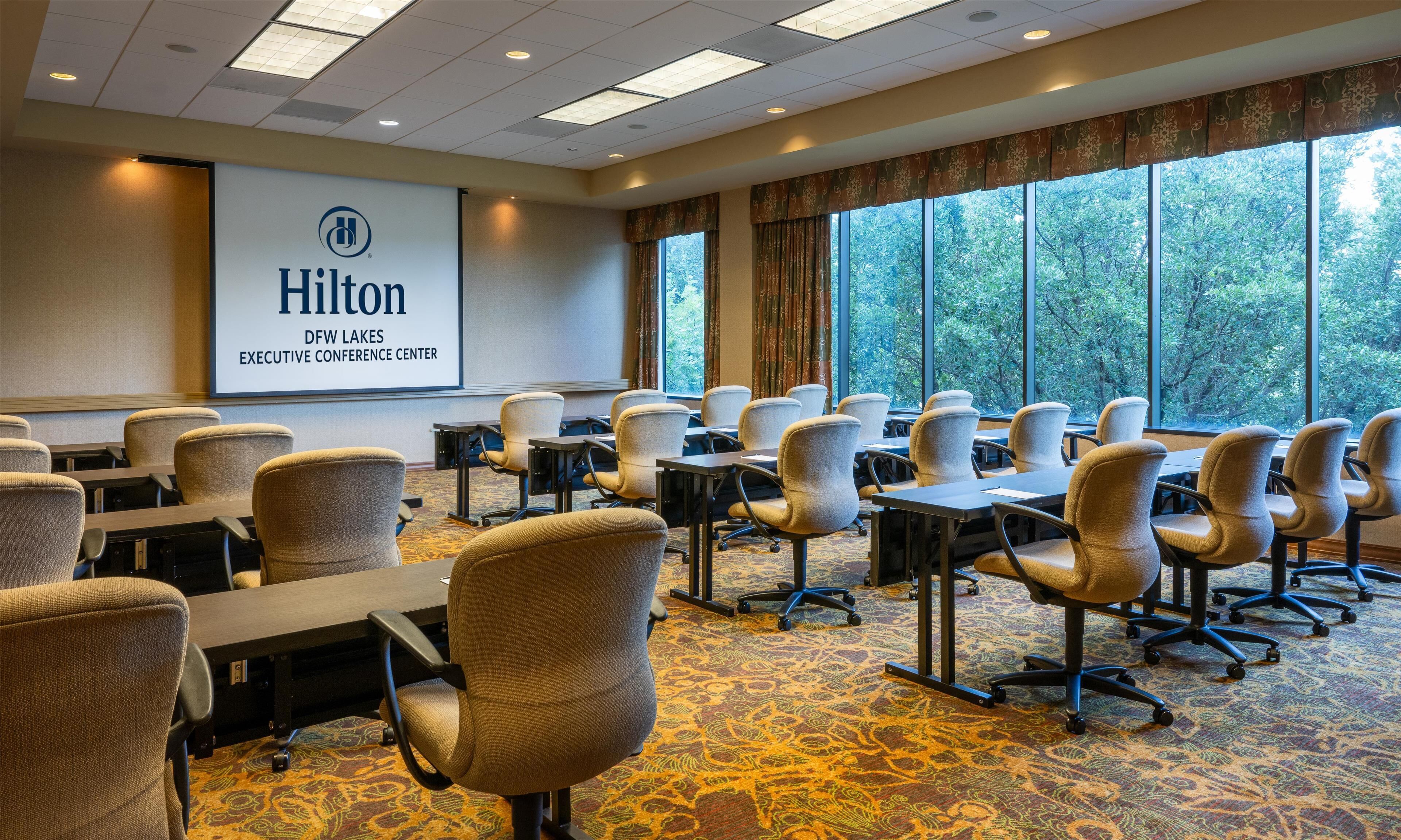 A conference room with rows of chairs, a projector screen displaying 'Hilton DFW Lakes Executive Conference Center,' and large windows with a view of trees.