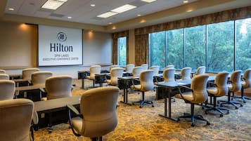 A conference room with rows of chairs, a projector screen displaying 'Hilton DFW Lakes Executive Conference Center,' and large windows with a view of trees.