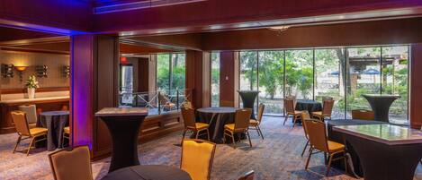 A hotel lobby with round tables and chairs, a reception area, and large windows offering a view of the outdoors.
