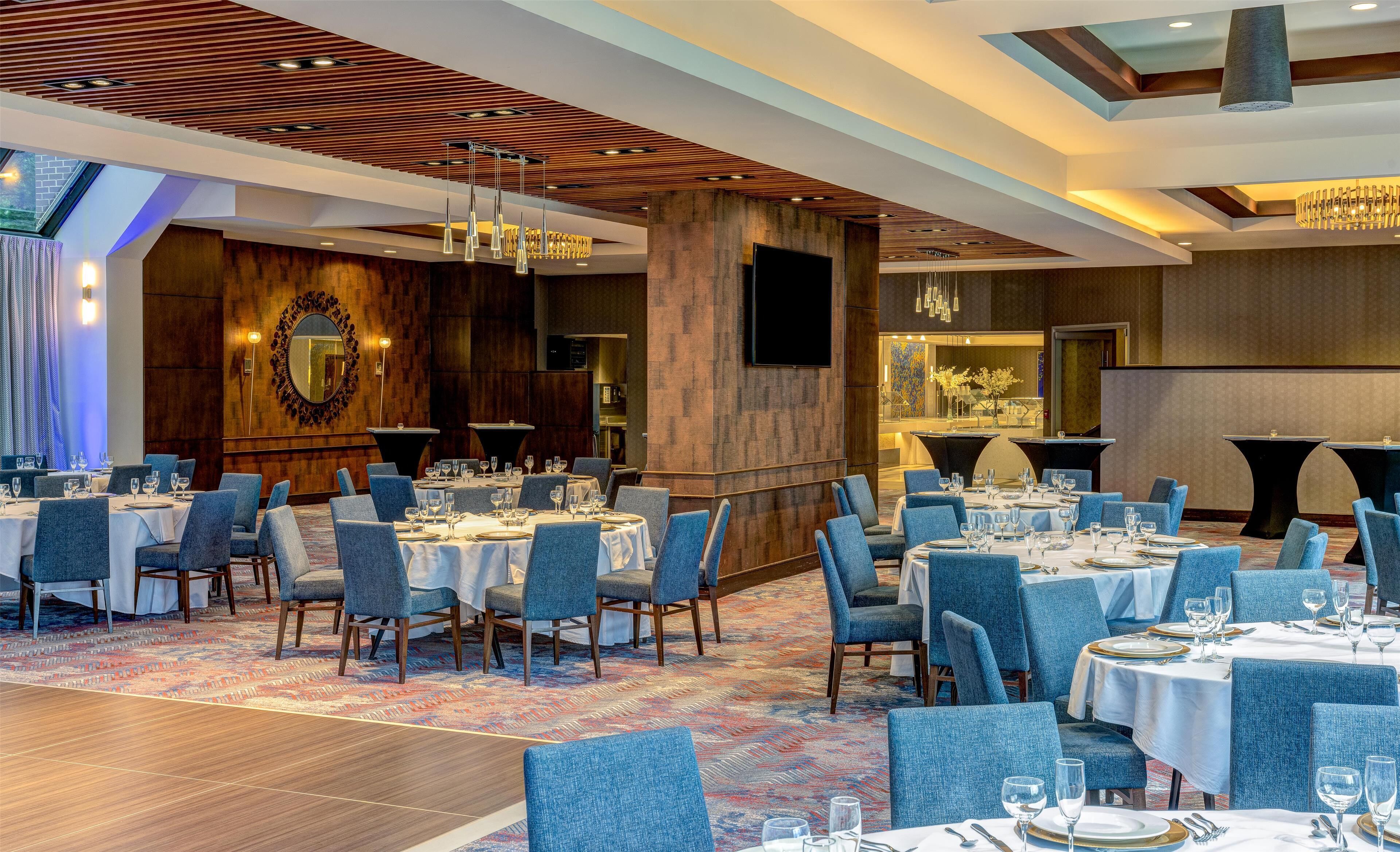 A well-lit dining area with multiple tables set for guests, featuring a modern ceiling design and a large mirror on the wall.