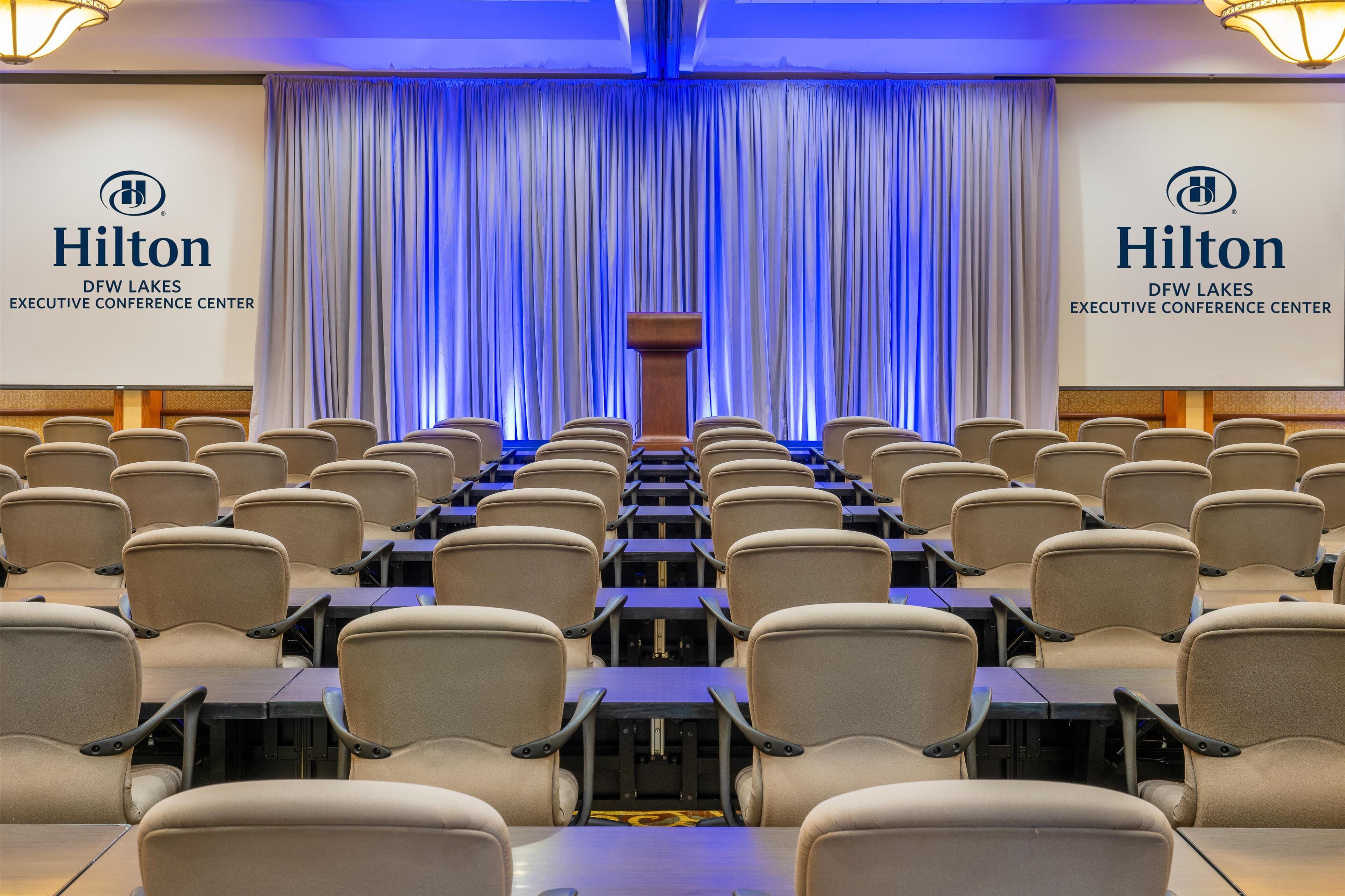 A conference room with rows of beige chairs and tables, a podium at the front, and a blue-lit curtain backdrop.