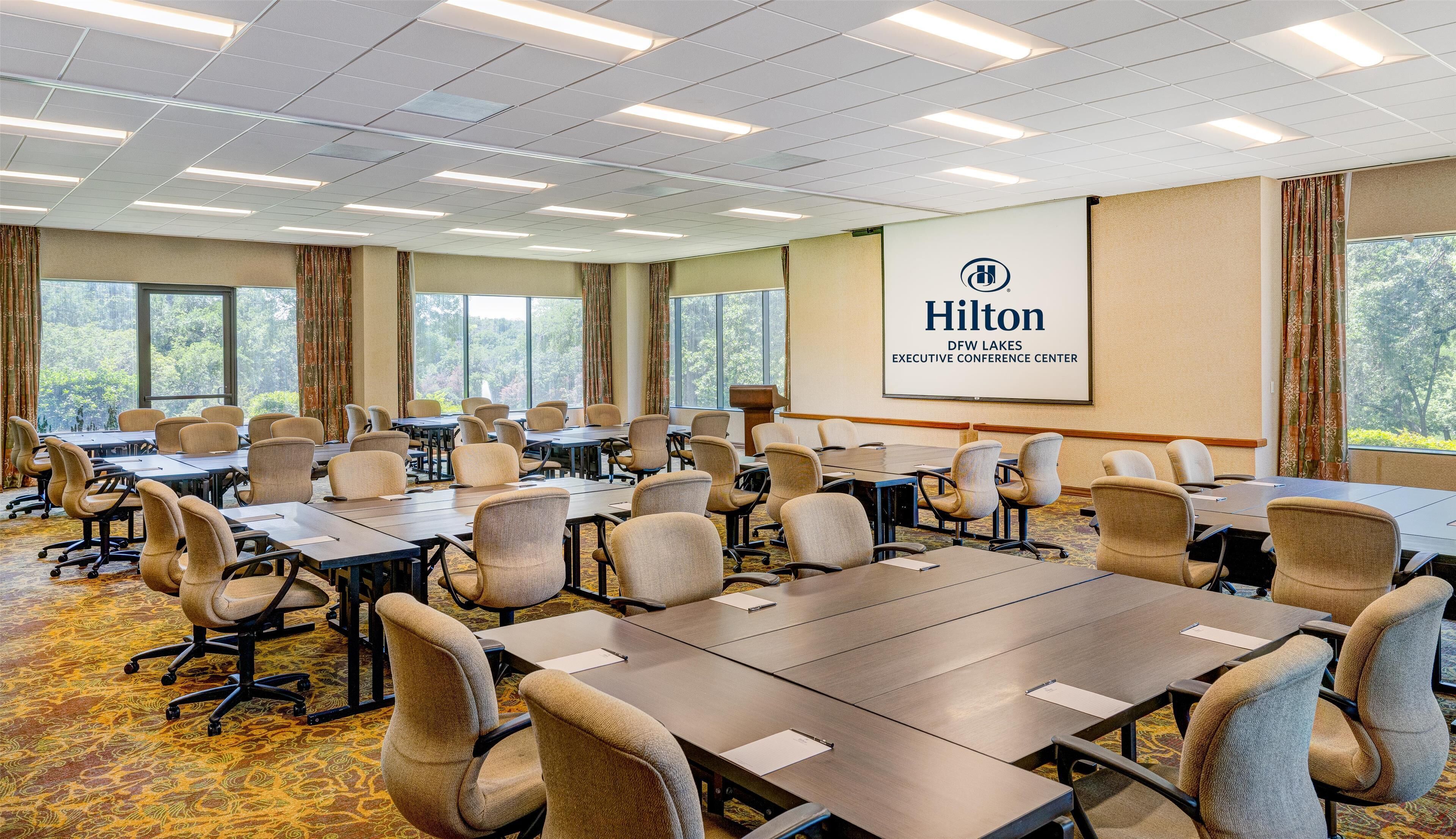 Een congreszaal met meerdere tafels en stoelen, grote ramen en een projectiescherm waarop het Hilton-logo staat.