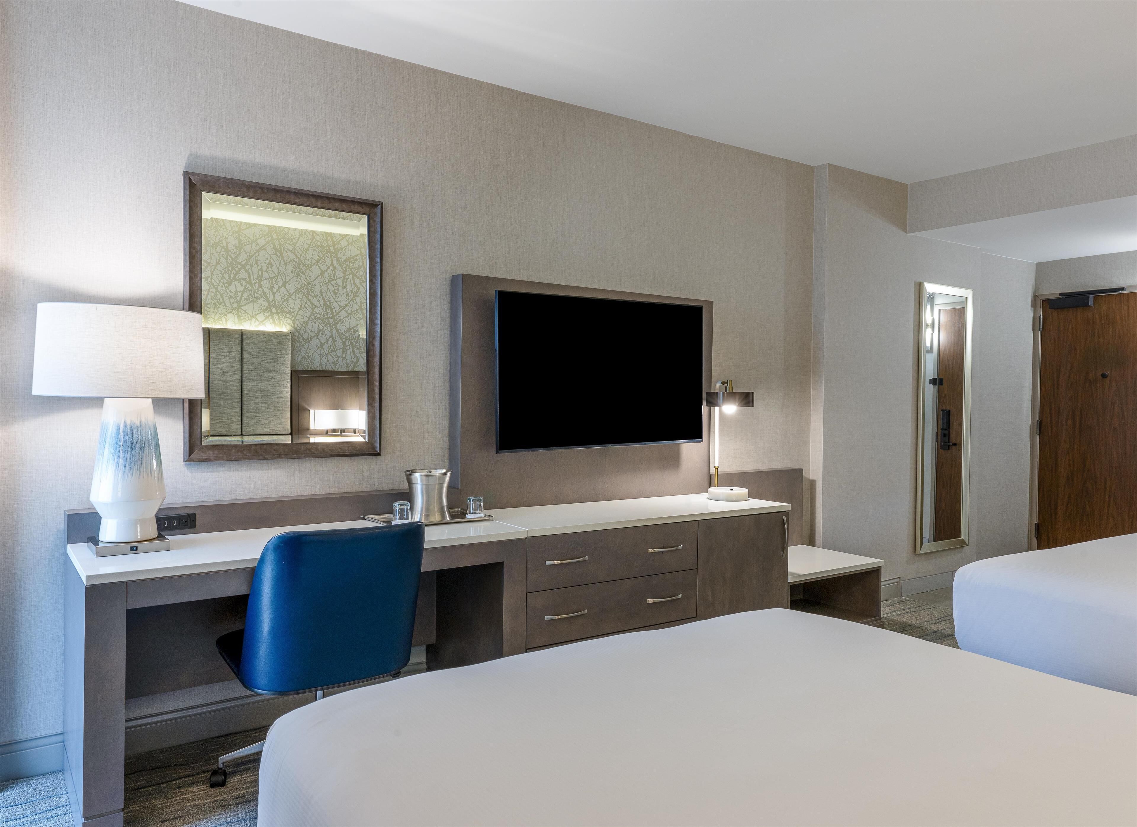 A hotel room with a desk, a chair, a bed, a television, and a mirror.