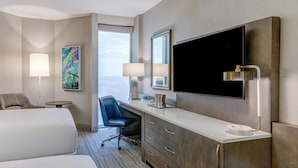 A hotel room with a large bed, a desk with a chair, a television, a large mirror, and a window with a view of the sky.