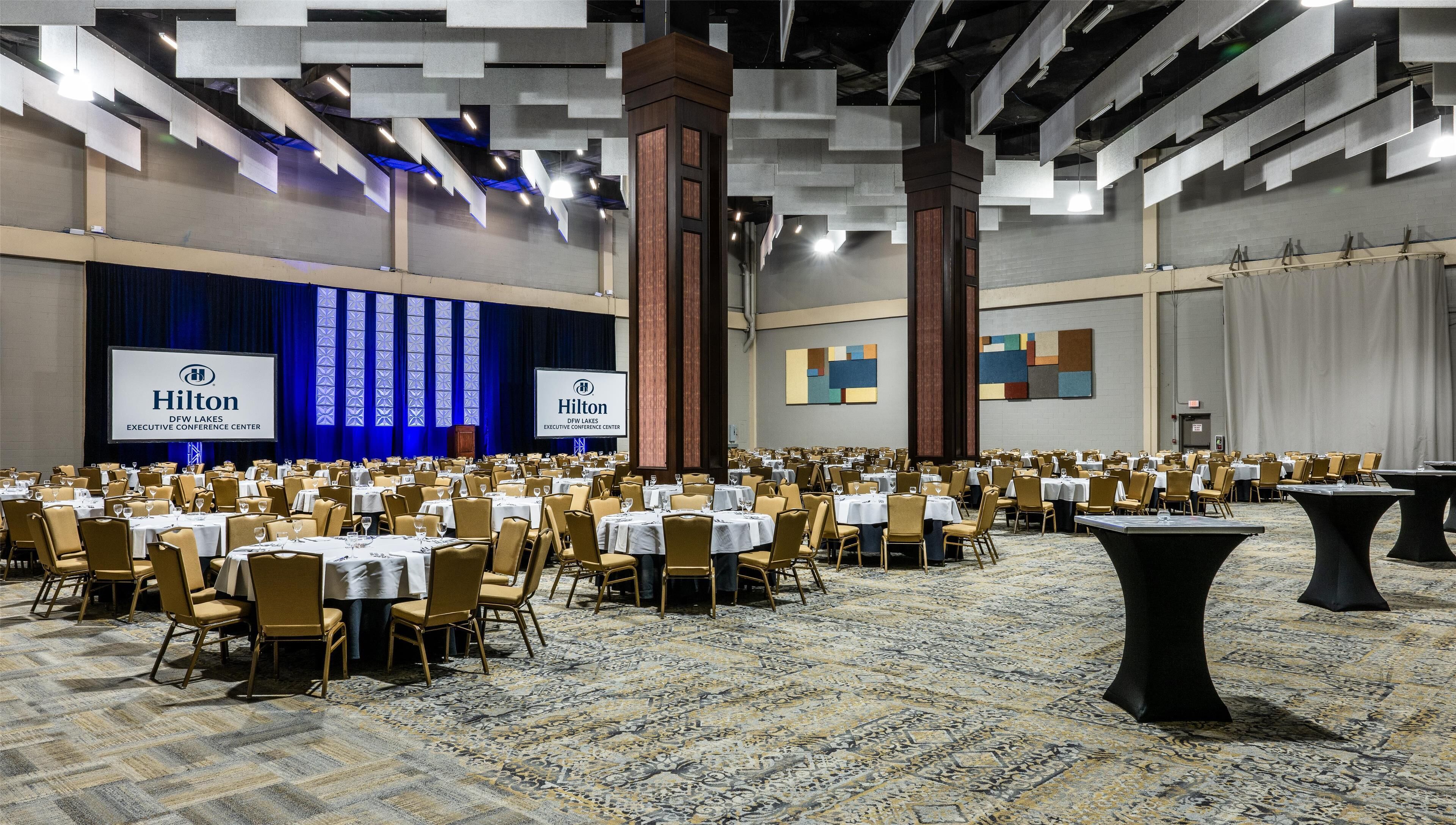 A large conference room with multiple tables and chairs, a stage with a presentation screen, and a carpeted floor.