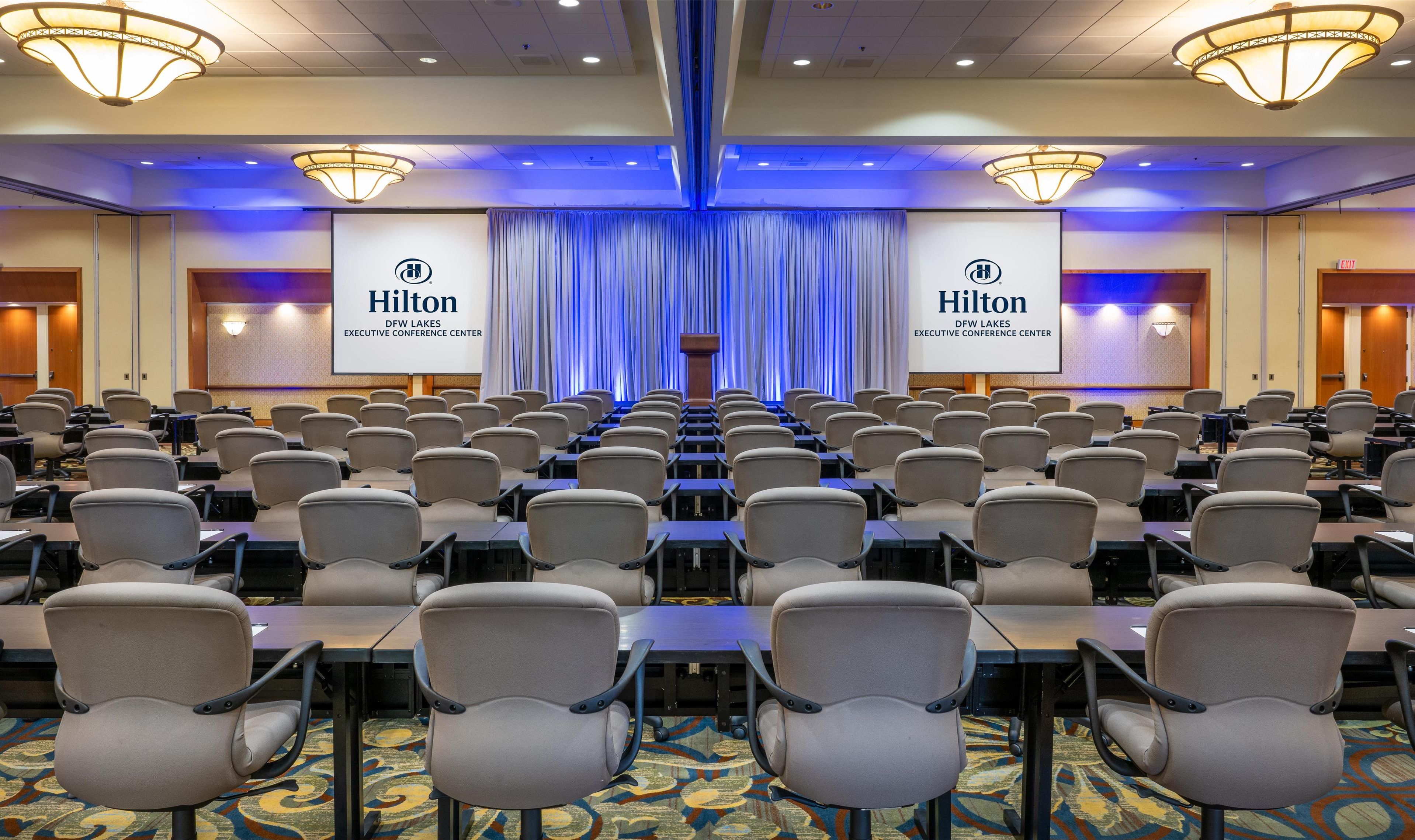 A conference room with rows of chairs and tables, a stage with a podium, and a large screen displaying the Hilton logo.