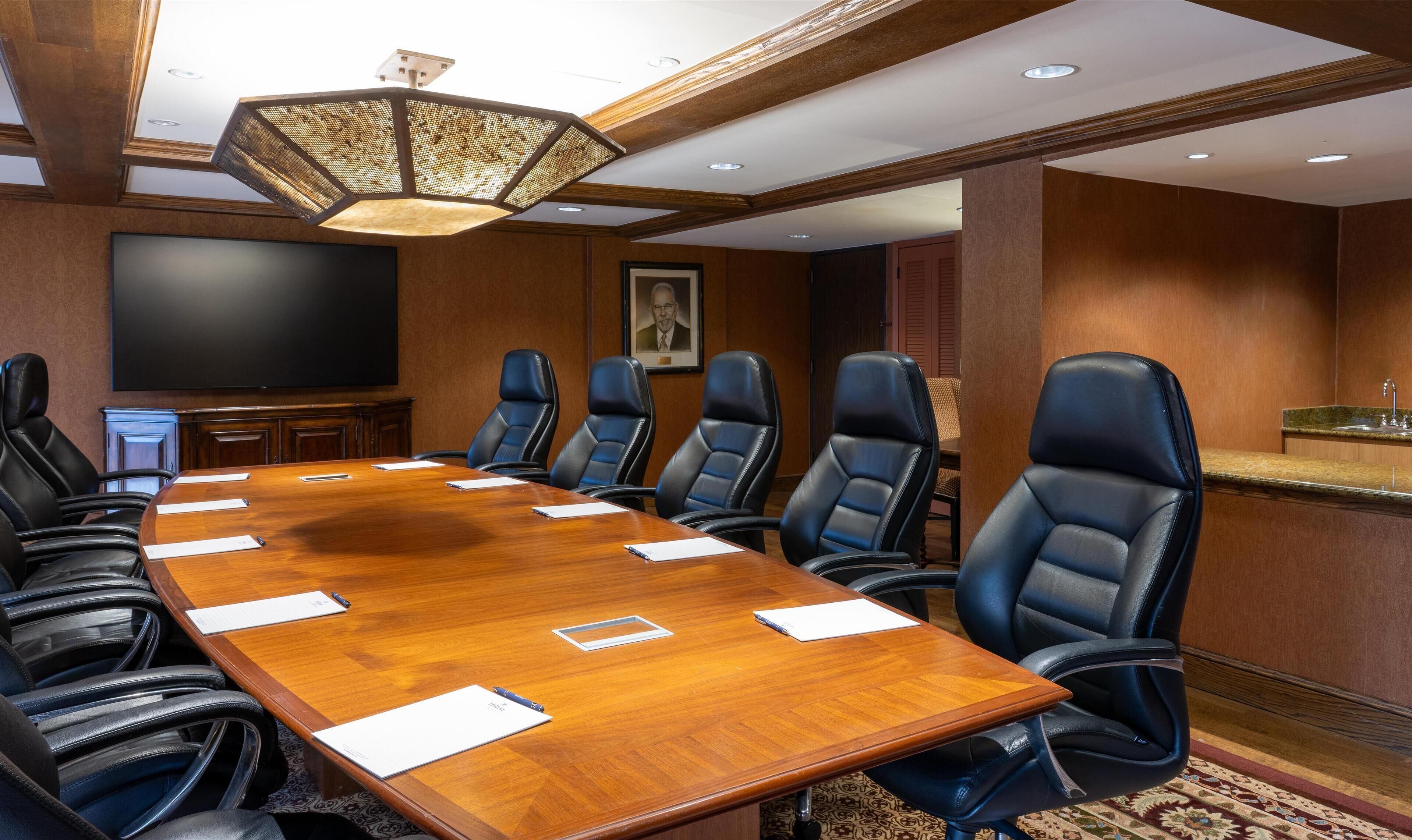 Une salle de conférence dotée d’une grande table en bois, de fauteuils en cuir noir, d’un téléviseur à écran plat et d’un lustre.