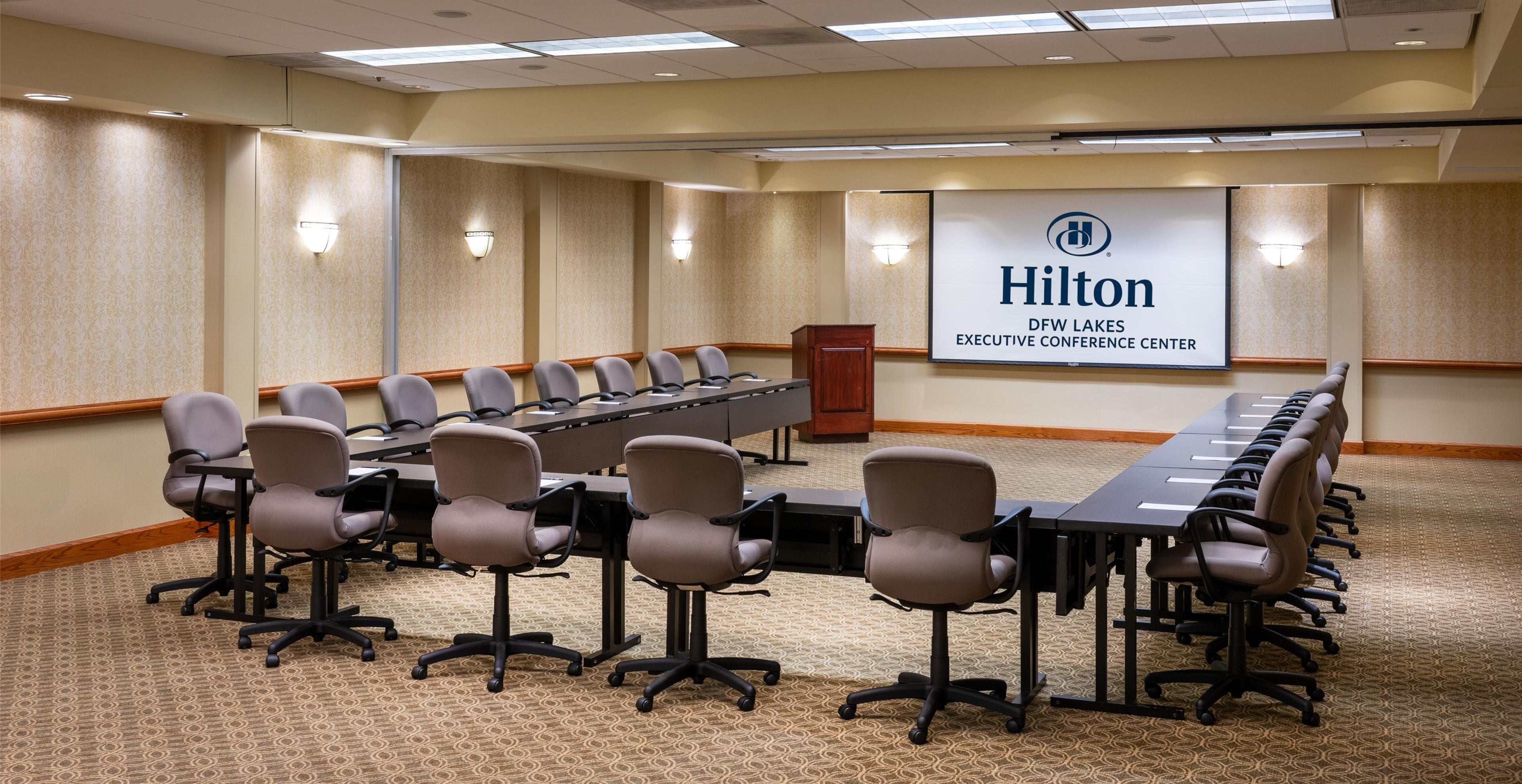 A conference room with a long table and multiple chairs, a podium, and a banner reading "Hilton DFW Lakes Executive Conference Center".