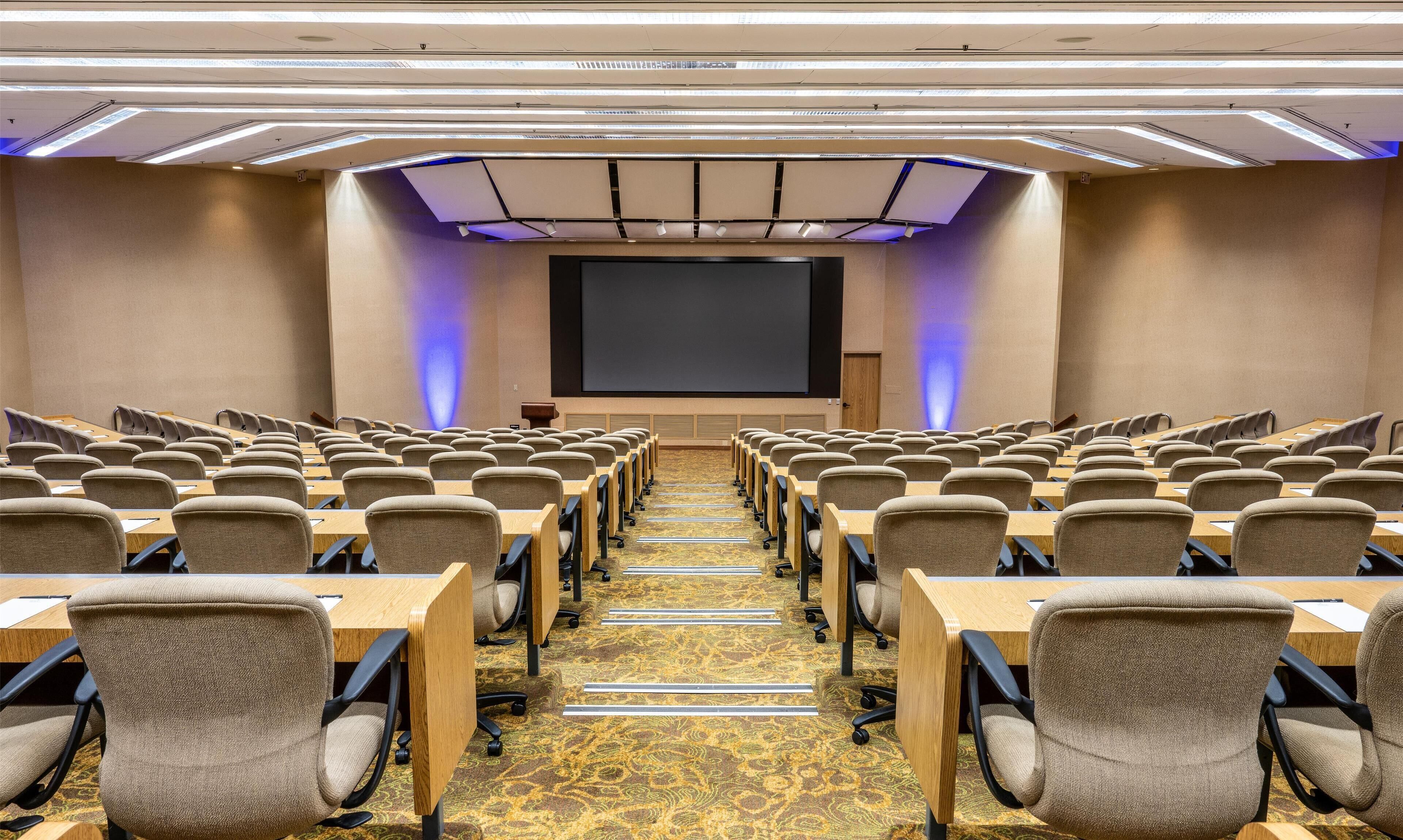 A conference room with rows of chairs and tables, a large screen at the front, and a carpeted floor.