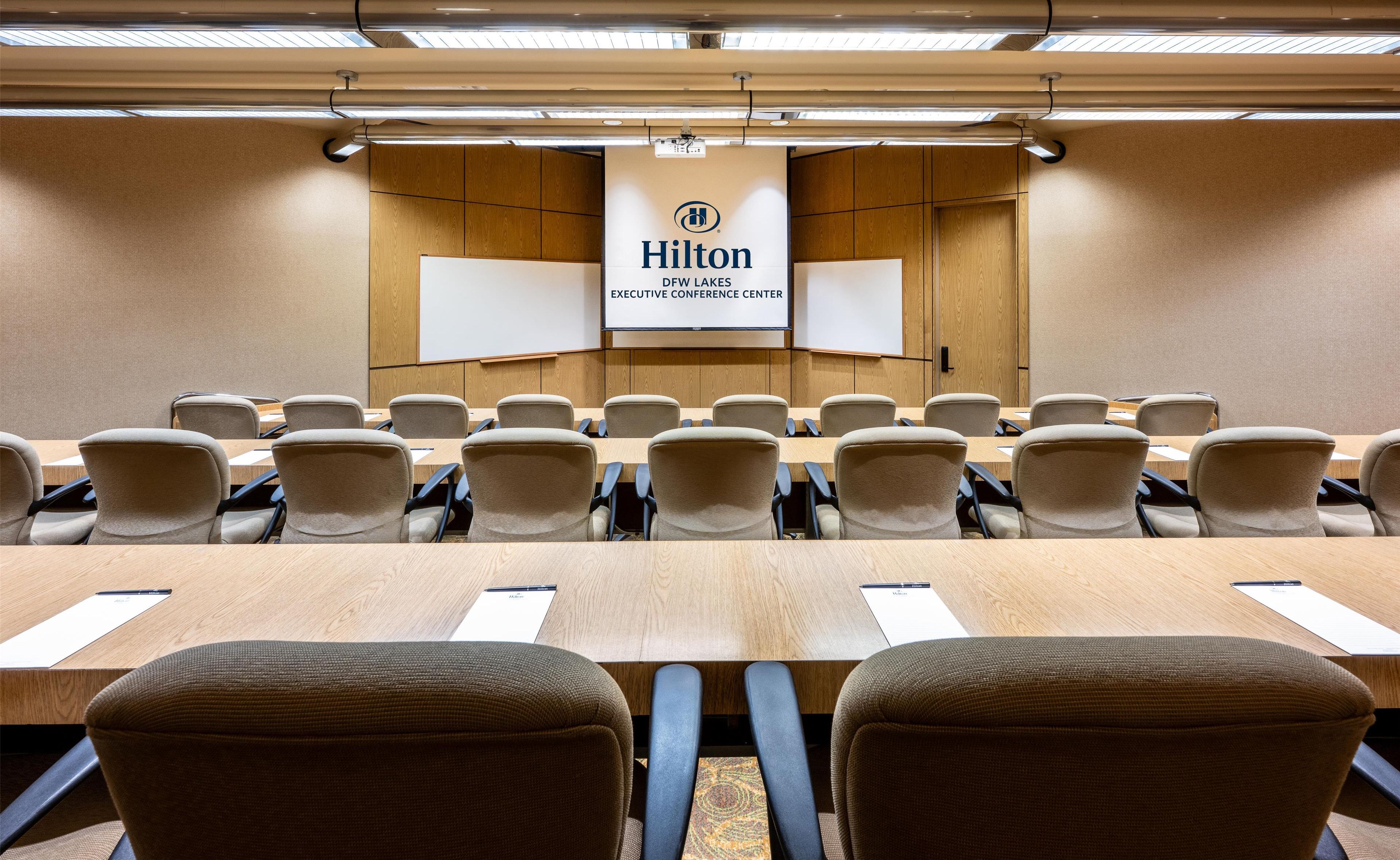 A conference room with a long table, multiple chairs, and a large screen displaying 'Hilton DEFW LAKES EXECUTIVE CONFERENCE CENTER'.