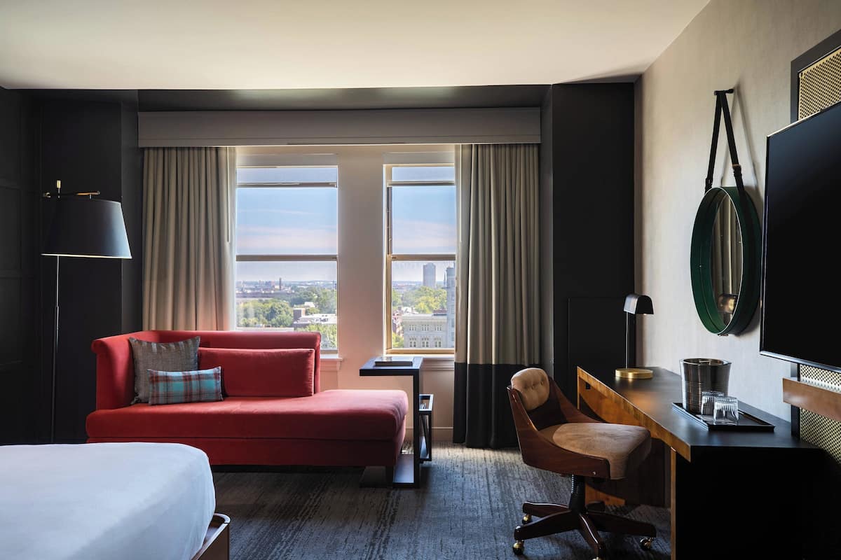Une chambre d’hôtel avec un canapé rouge, un bureau en bois, une chaise, un lit et une grande fenêtre donnant sur la ville.